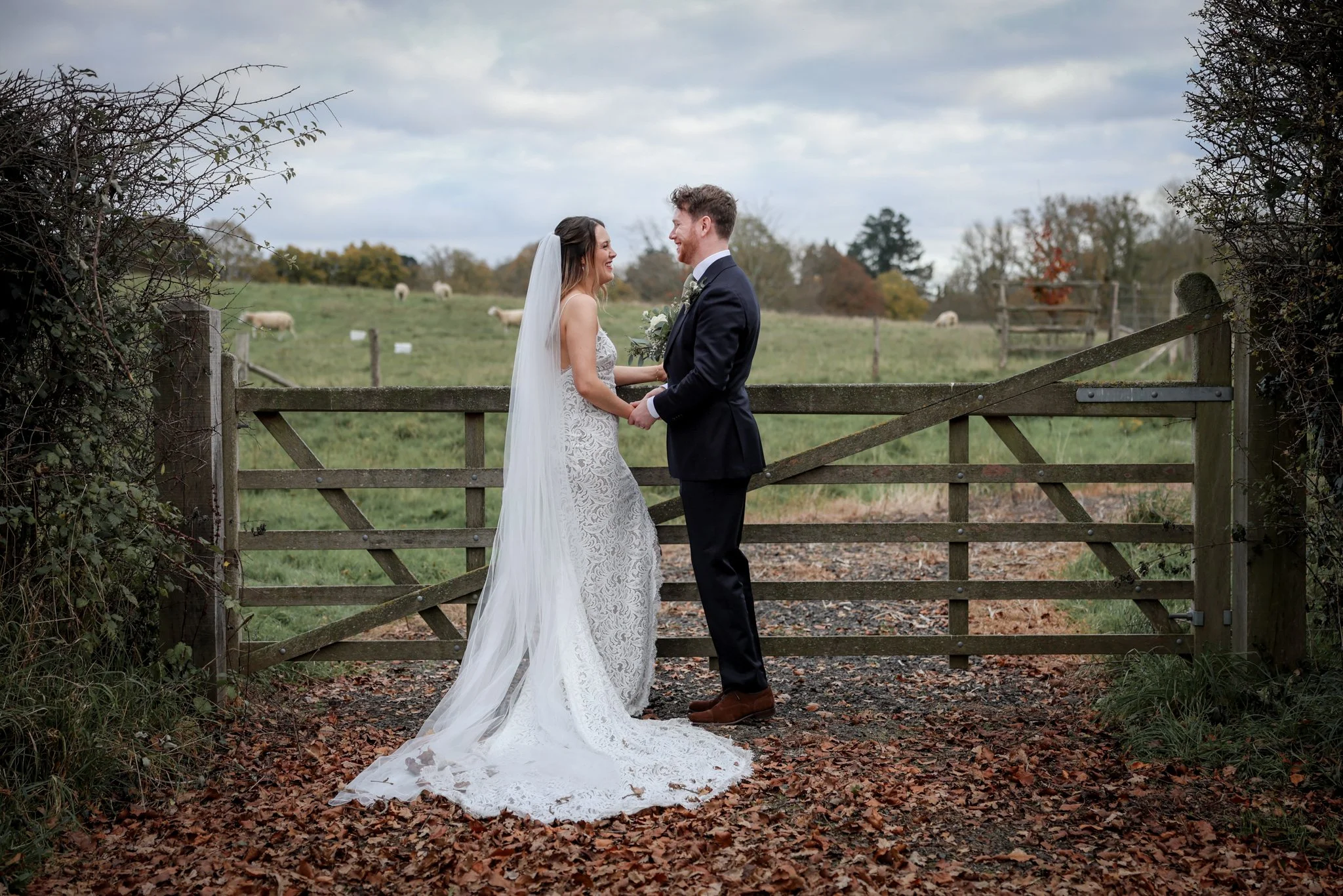 A bride and groom holding hands and smiling at each other in front of a wooden gate, outdoors in a rural setting with a field and sheep in the background, overcast sky.