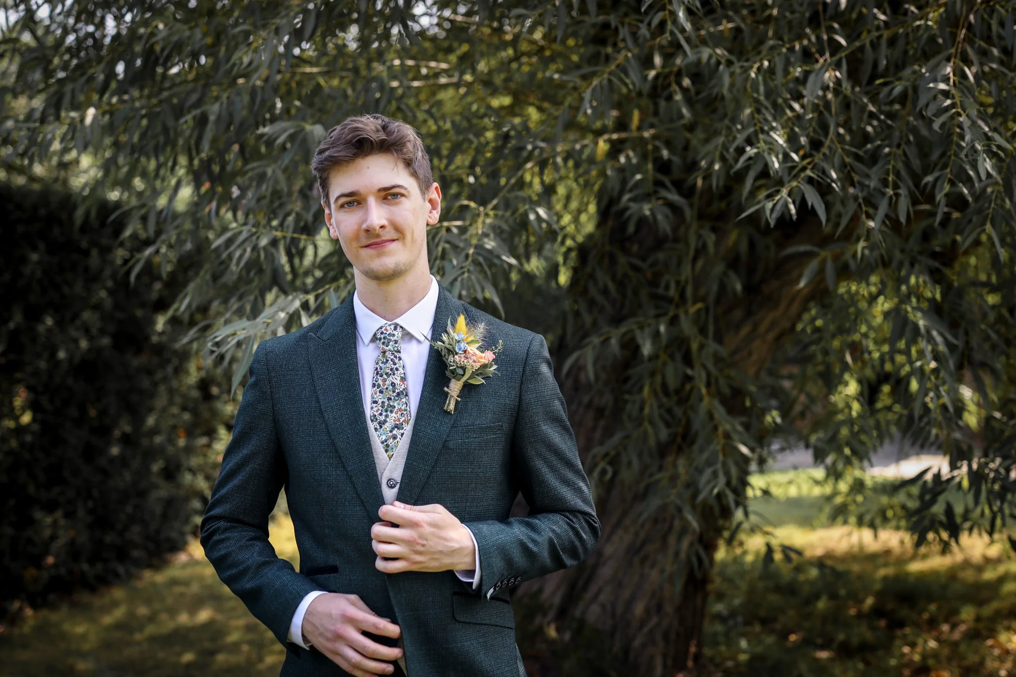 Young man in a suit with a floral boutonniere standing outdoors near a large leafy tree.