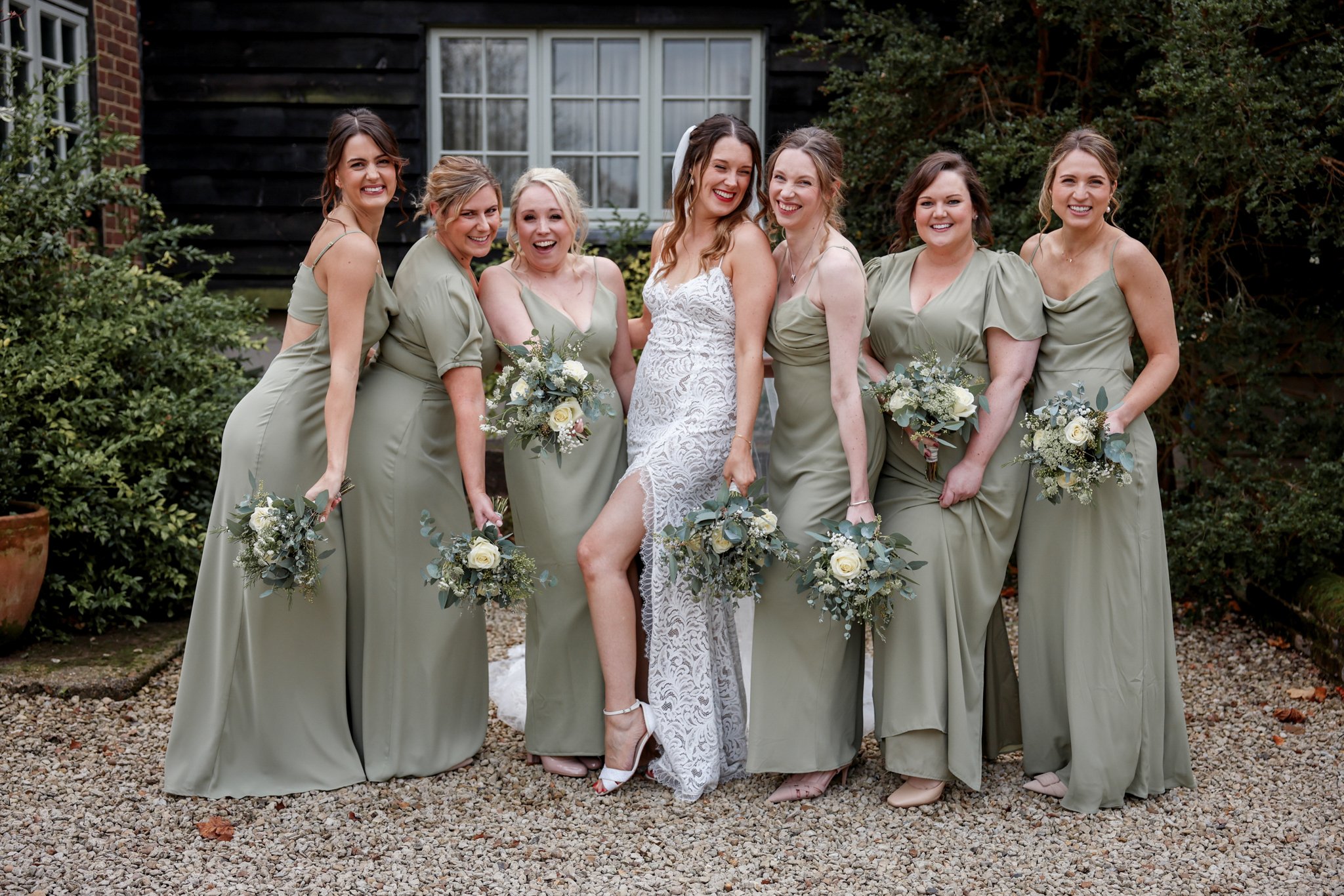 A group of seven women, including a bride and six bridesmaids, standing outdoors in front of a black house with a window. The women are dressed in matching sage green dresses, and the bride is wearing a white lace dress with a veil. They are smiling,