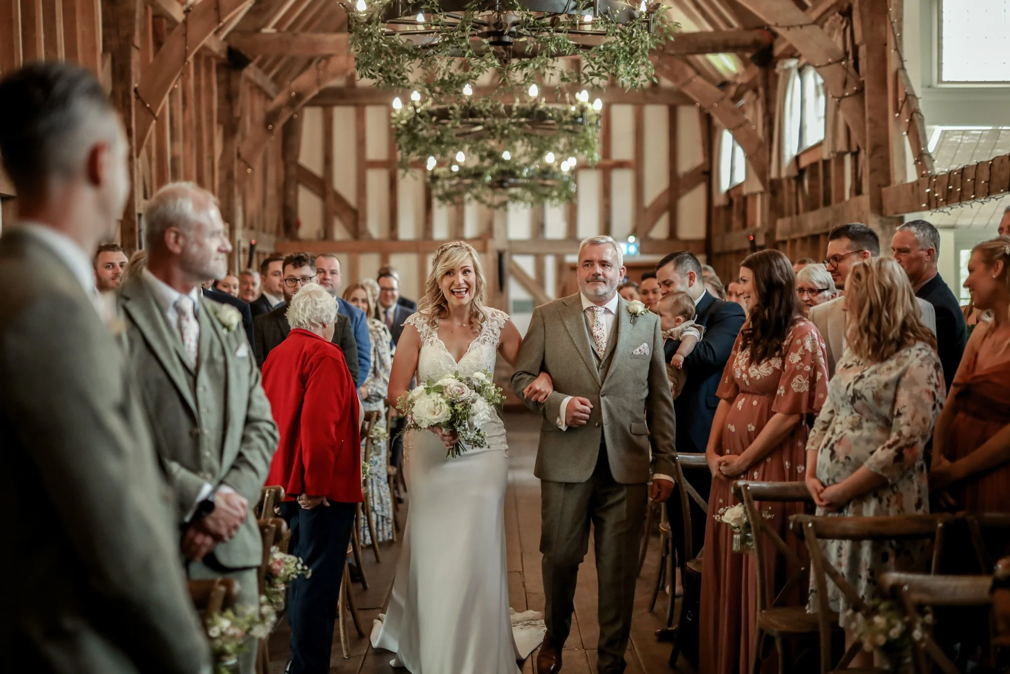 A bride walking down the aisle with her father at a rustic wedding in a barn with wooden beams, decorated with greenery and string lights.