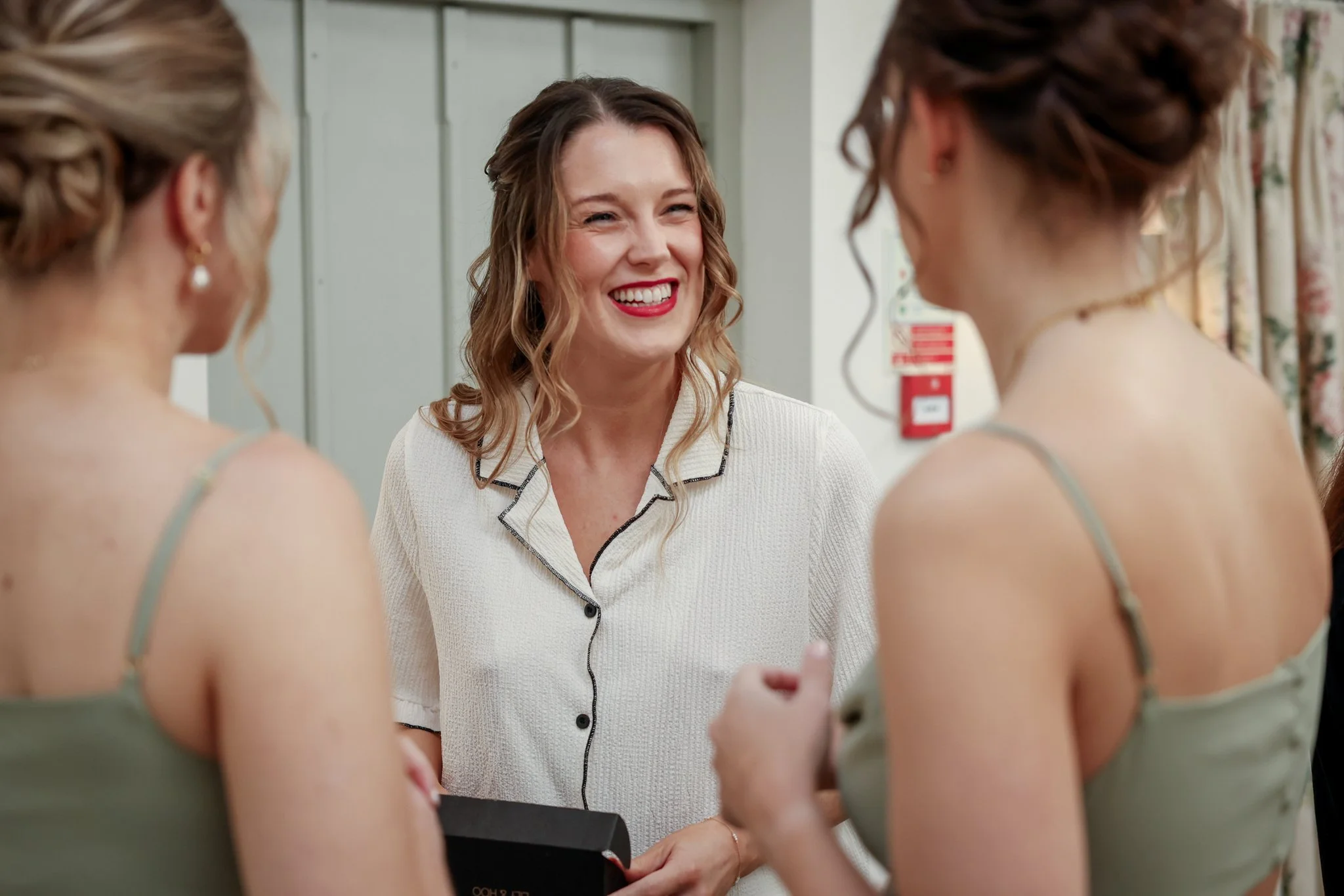 A woman with wavy brown hair and a white shirt smiling while talking to two women dressed in green tops with low-cut straps, in a room with light-colored walls and floral curtains.