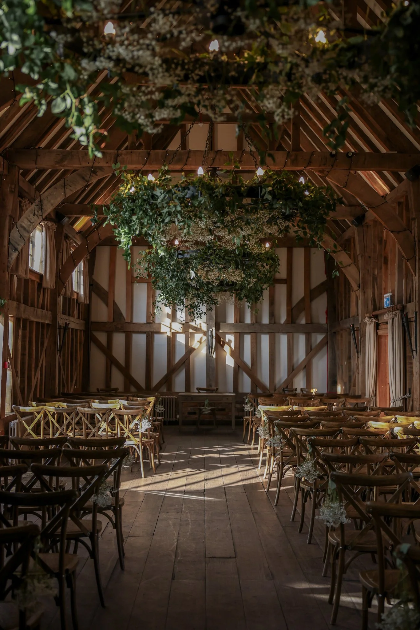 Interior of a rustic wooden barn decorated for a wedding or event, with hanging greenery and string lights, arranged with chairs and tables.