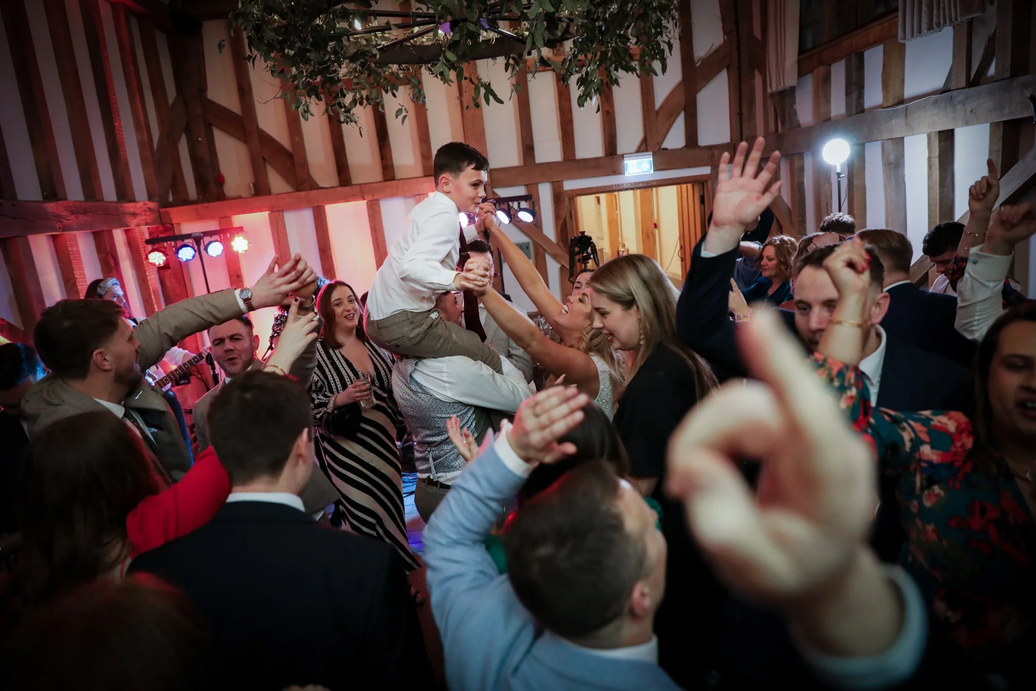 People celebrating at a wedding reception with a young boy being lifted on top of adults' hands, surrounded by dancing guests and colorful lights.