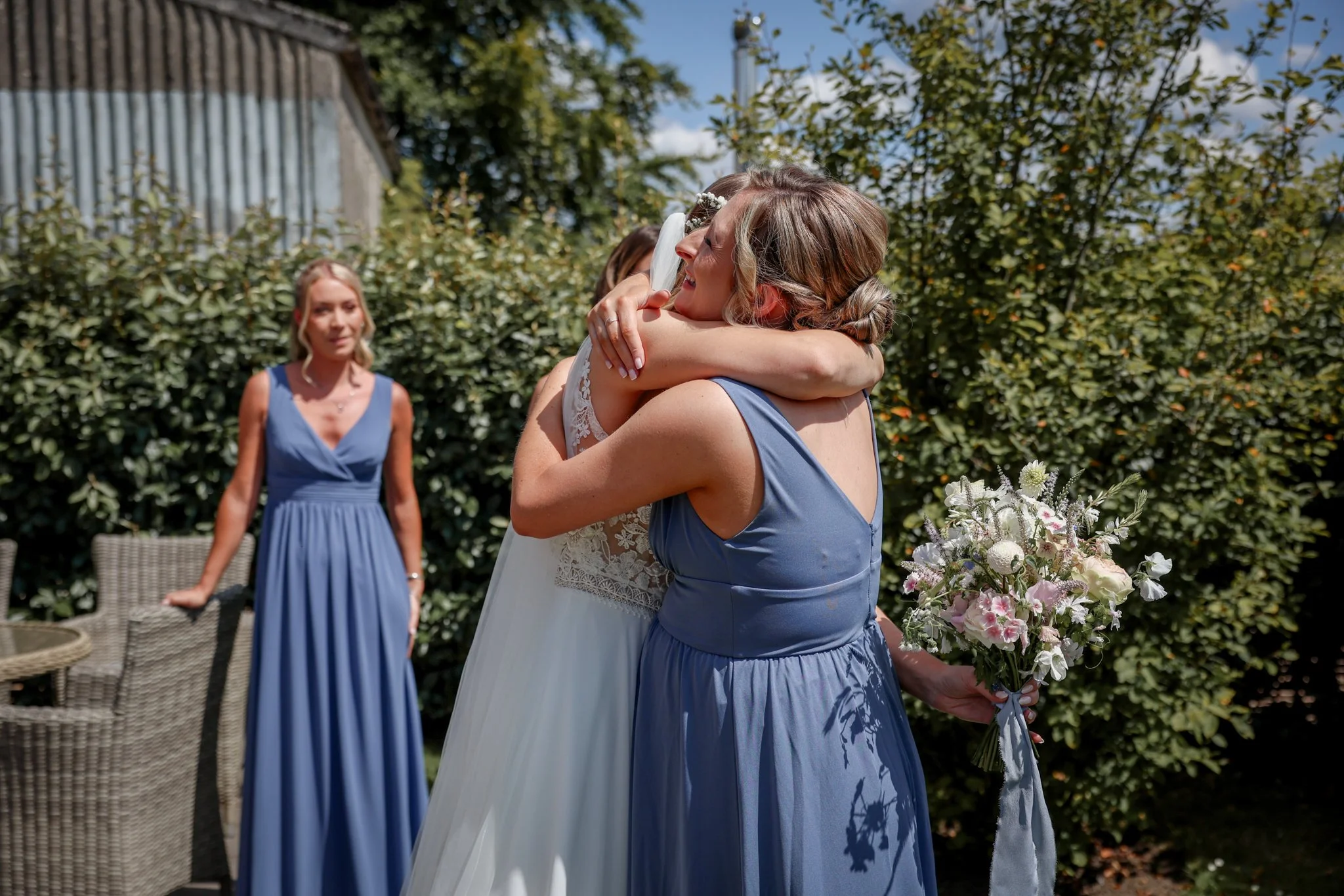 A woman in a blue dress hugging another woman in a wedding dress, standing outdoors with greenery and a blue sky in the background. A third woman in a similar blue dress is in the background observing.