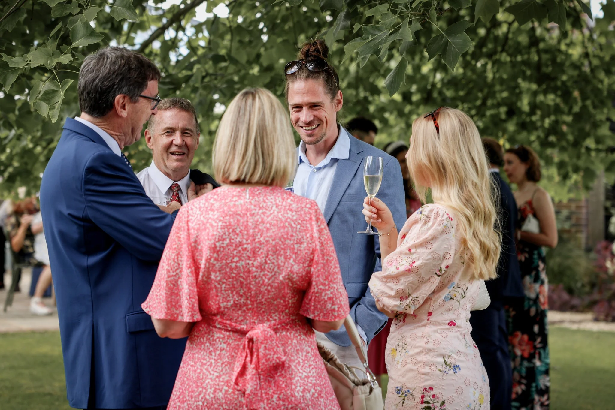 Group of five people smiling and talking at an outdoor event under trees, one woman holding a glass of champagne.