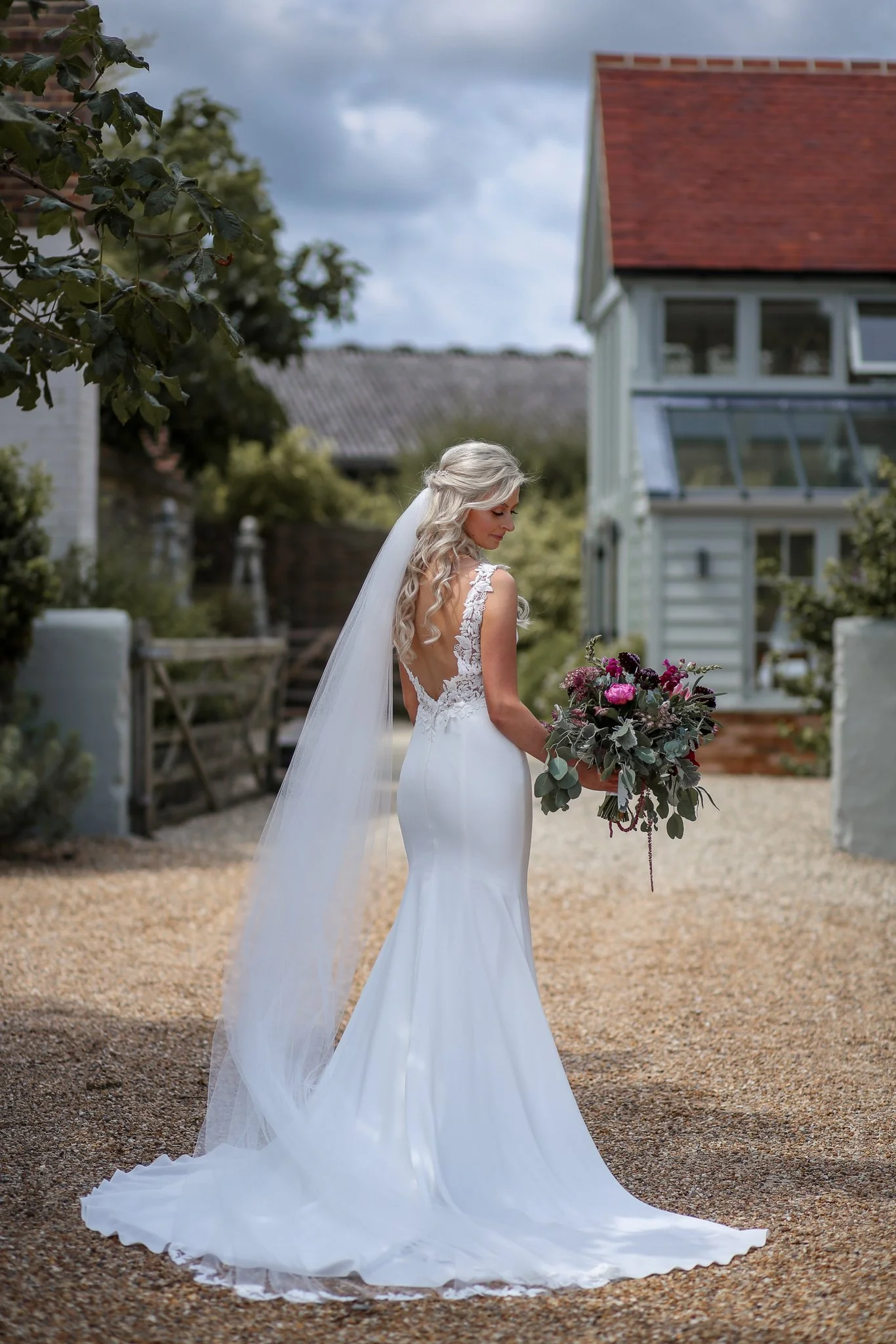 A bride with long, curly blonde hair in a white wedding gown with lace details and a long veil, holding a bouquet of pink, purple, and green flowers, standing outdoors with a house and trees in the background on a cloudy day.