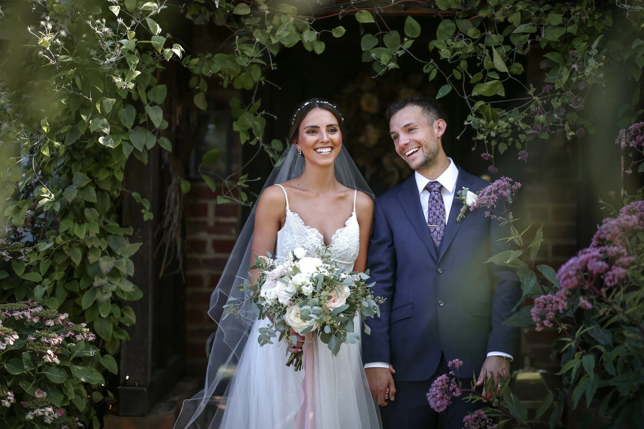 A bride and groom standing together outdoors, surrounded by green foliage and purple flowers, smiling happily.
