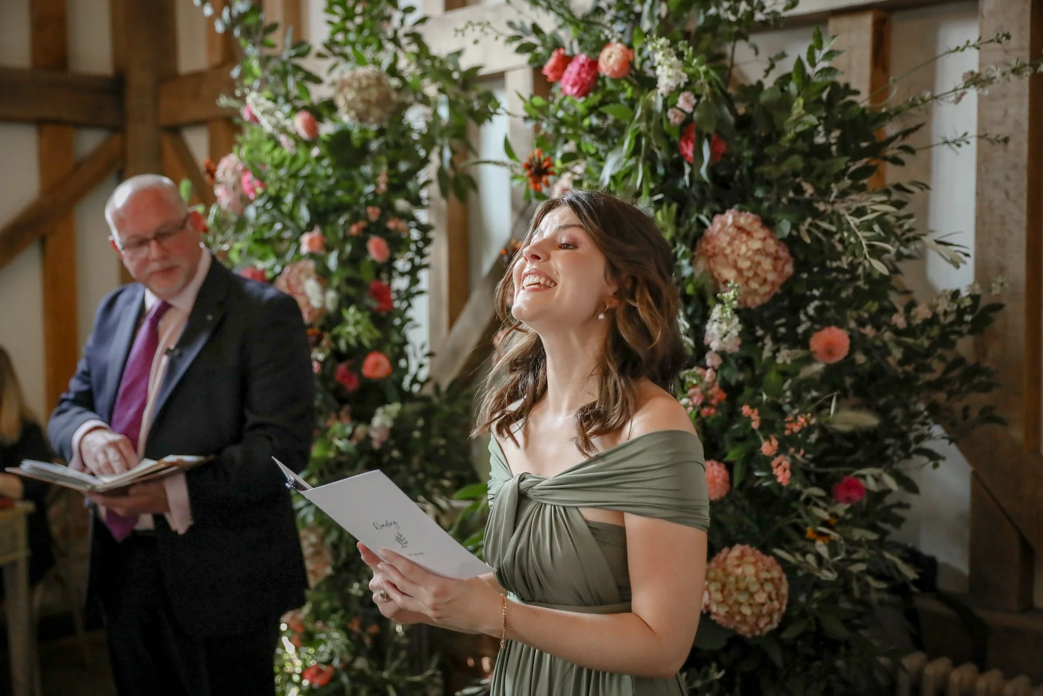 A woman smiling and holding a booklet, with a man reading a book beside her, in front of a large floral arrangement at an indoor event.