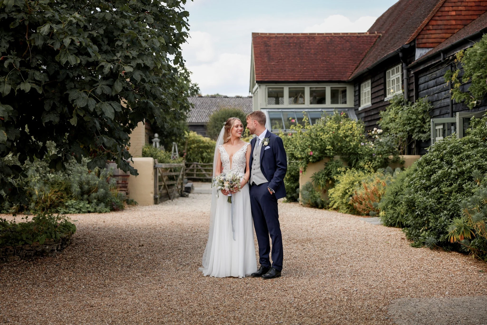 A bride and groom smiling and standing close together outdoors in a garden setting, with rustic houses and lush greenery in the background.