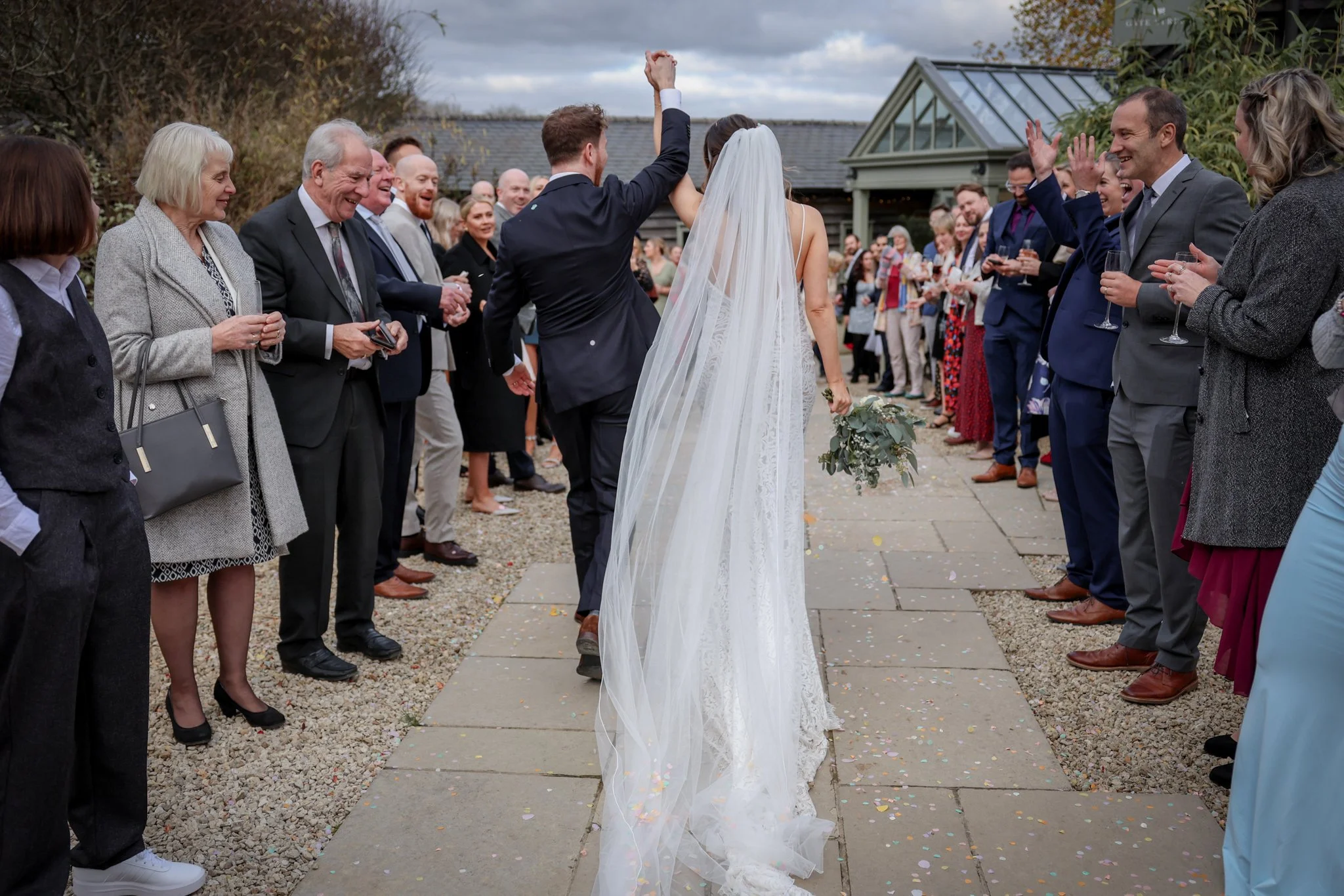 A bride and groom stand together with the groom raising his arm in celebration, surrounded by smiling friends and family at an outdoor wedding ceremony.