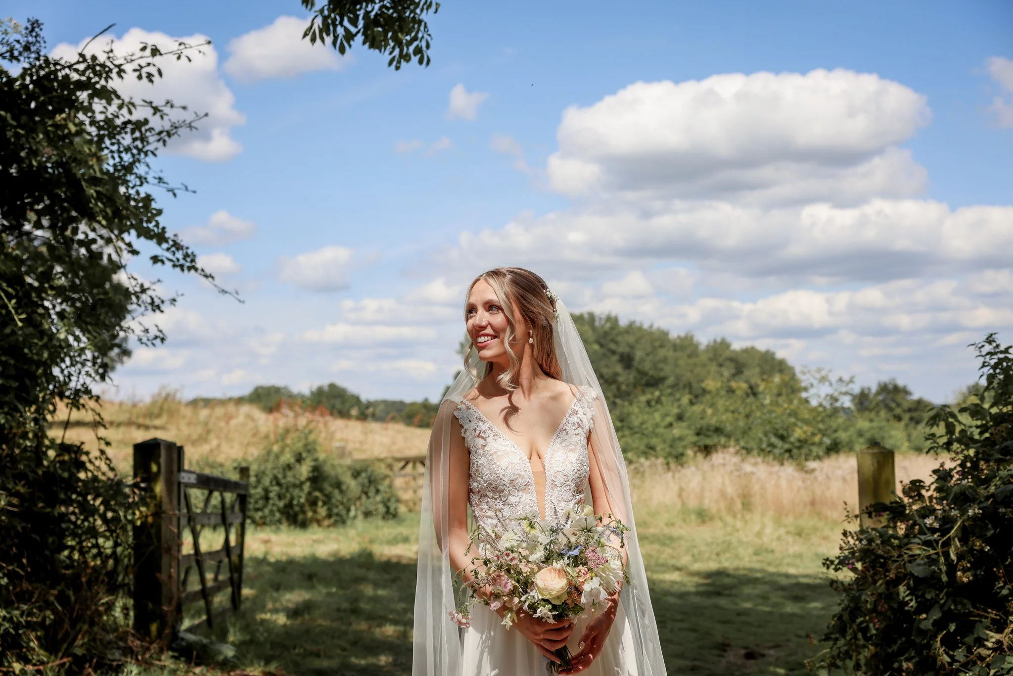 A bride standing outdoors on a sunny day, holding a bouquet of flowers, wearing a lace wedding dress and veil, smiling with trees and a blue sky with clouds in the background.