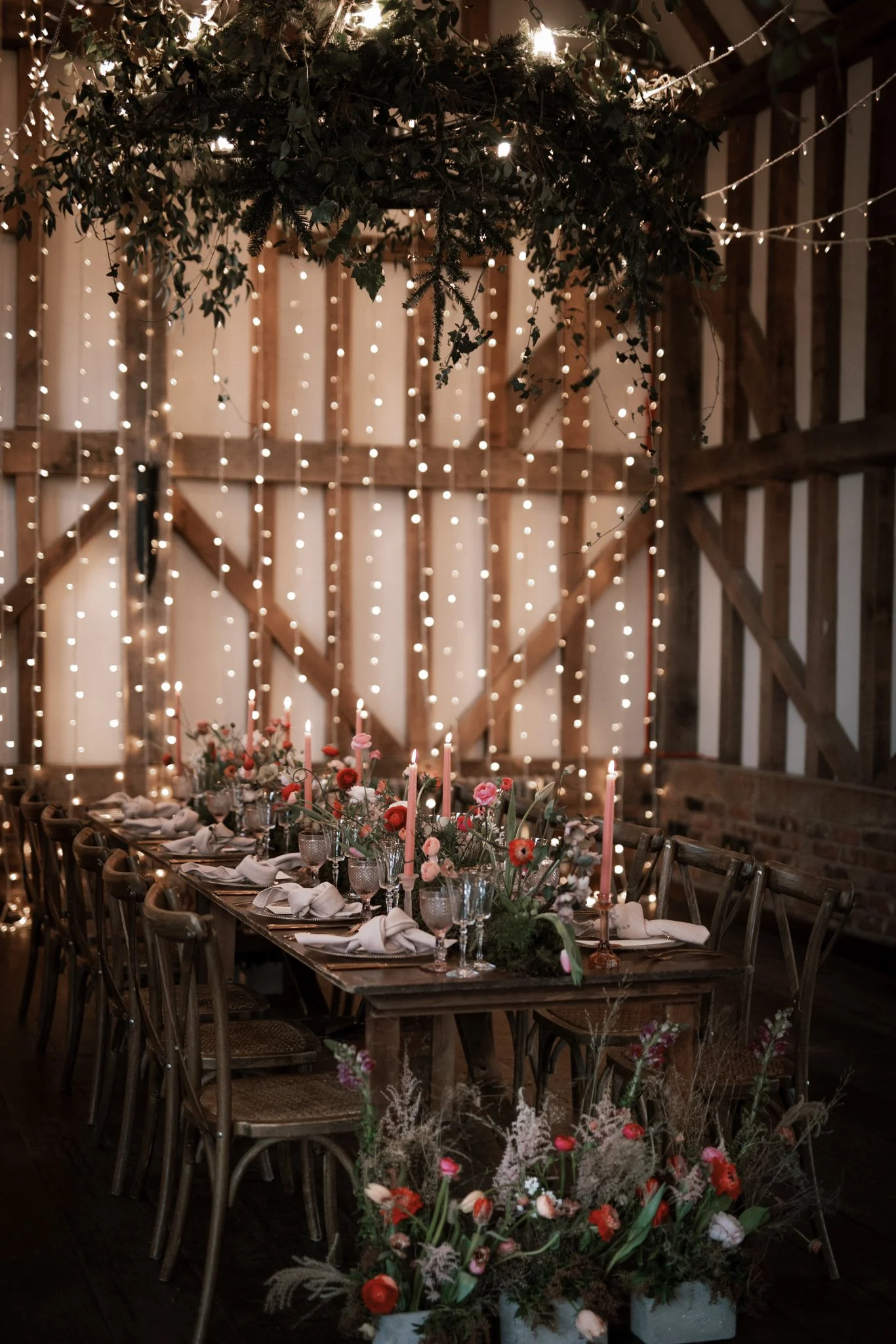 A rustic indoor wedding reception setup with a long wooden table decorated with flowers, candles, and tableware. Fairy lights hang above, and a greenery chandelier is suspended from the ceiling.