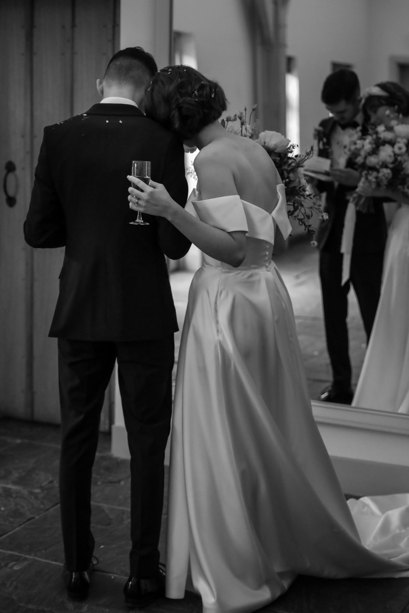 Black and white photo of a couple at their wedding, embracing with heads touching, the woman holding a glass of wine, in a room with a mirror and other people celebrating.