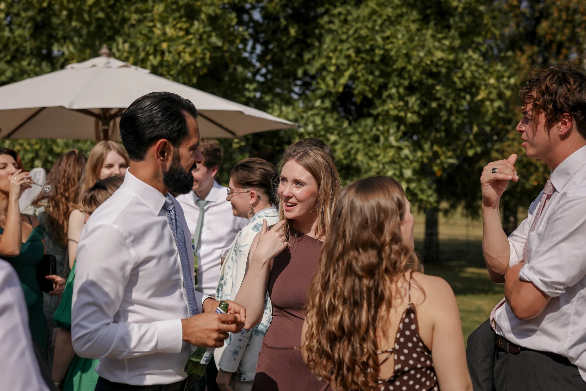 Group of people socializing outdoors under a large umbrella surrounded by trees, dressed in semi-formal attire.