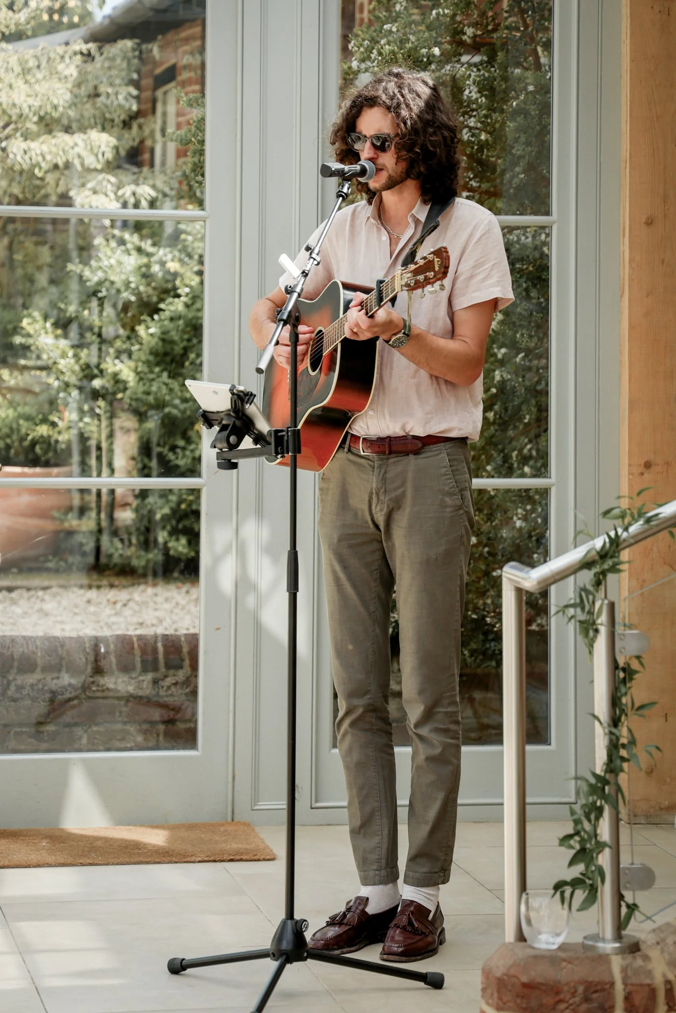A man with curly hair, sunglasses, and a beige shirt is playing an acoustic guitar and singing into a microphone. He is standing indoors near a glass door with greenery outside.