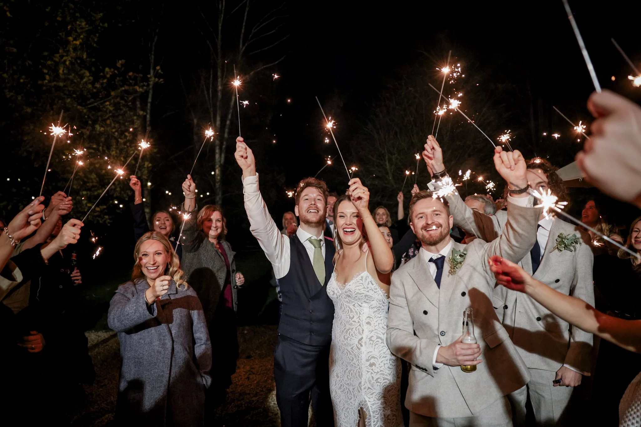 Group of people celebrating at night with sparklers, smiling and dressed in formal attire, possibly at a wedding or special event.
