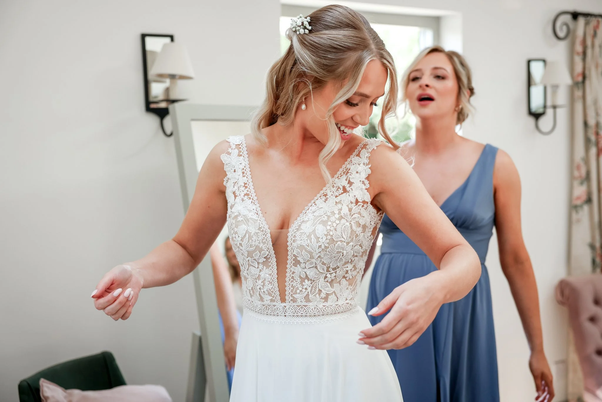 A bride with blonde hair in a lace wedding dress smiling as she adjusts her gown, with a woman in a blue dress assisting her in a bright, decorated room.
