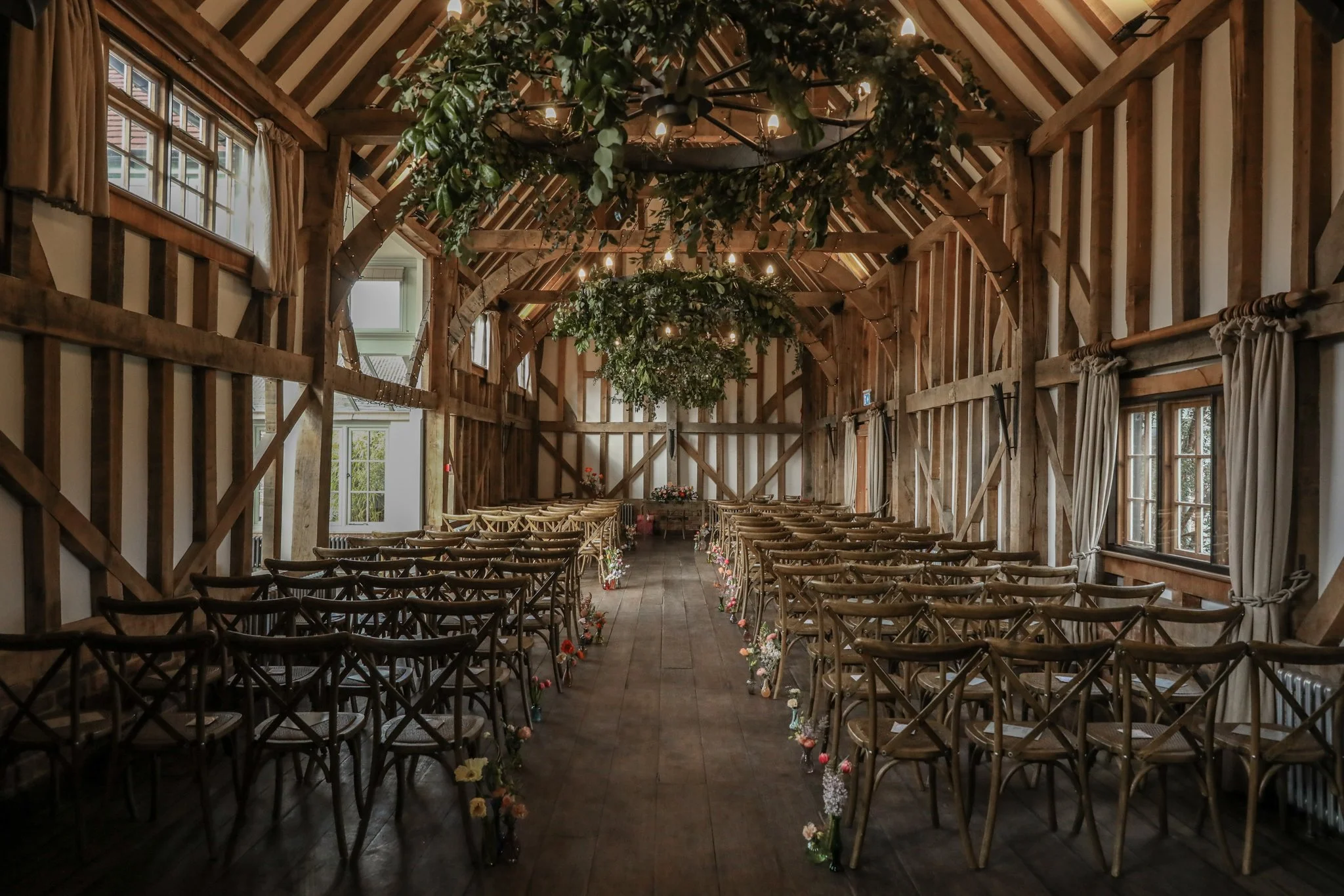 Indoor rustic wedding ceremony space with wooden beams, rows of chairs, floral decorations, and greenery arching overhead.