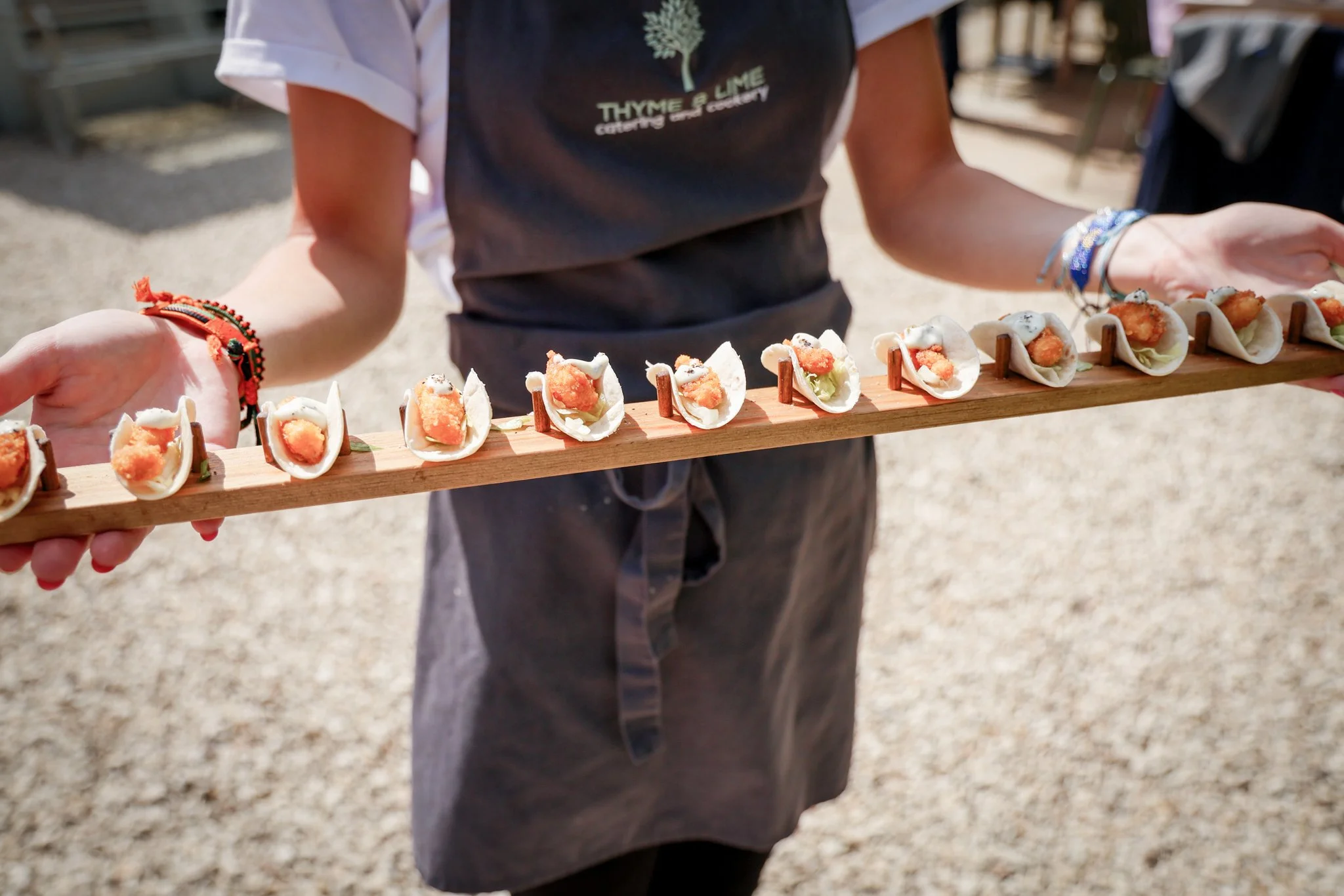 Person holding a wooden tray with small tacos filled with fried fish, lettuce, and white sauce, wearing bracelets, in an outdoor setting.