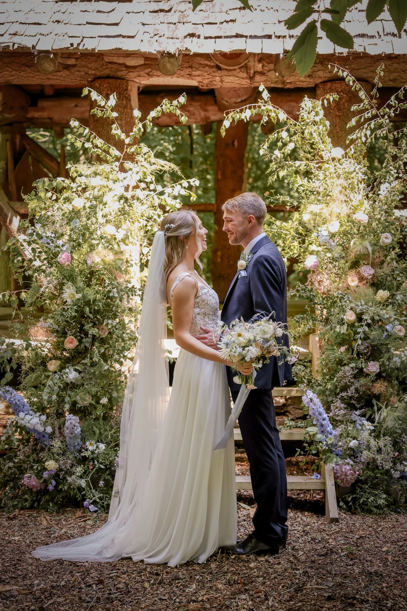 A bride and groom standing face to face at their outdoor wedding ceremony, surrounded by flowers and greenery, holding hands and smiling at each other.