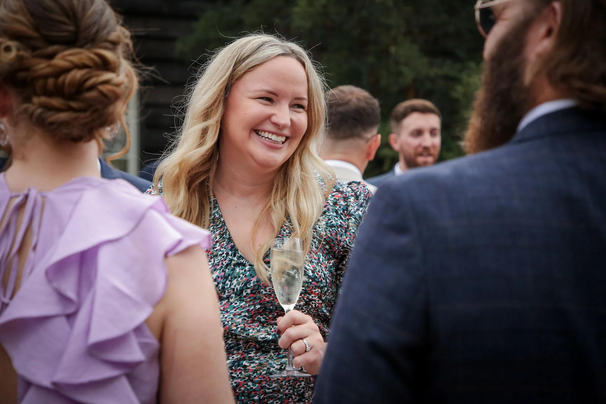 A group of people at an outdoor event, with a woman smiling and holding a glass of champagne, surrounded by others dressed in formal attire.