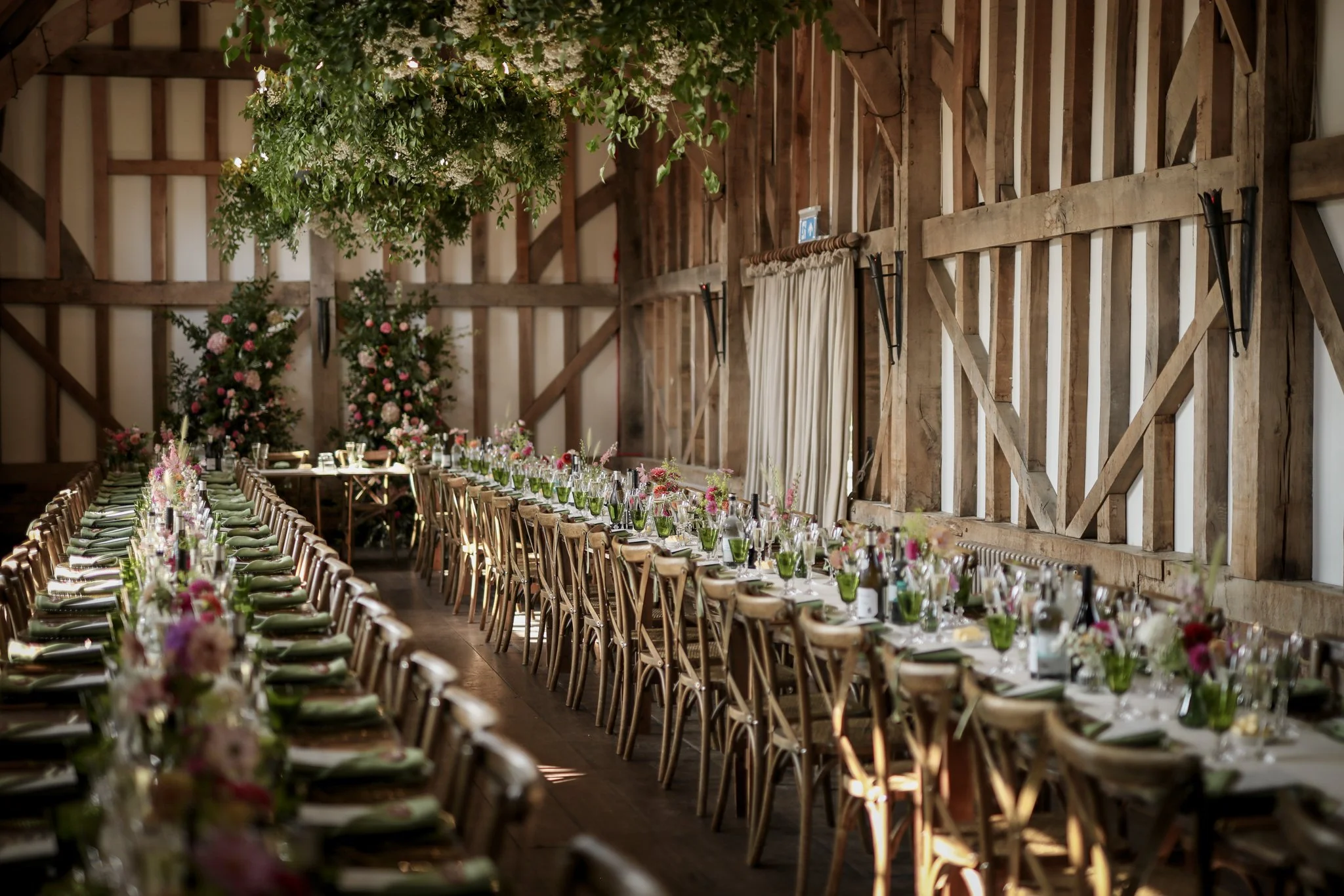 Long dining table set with flowers and tableware inside a rustic barn with wooden walls and hanging greenery.