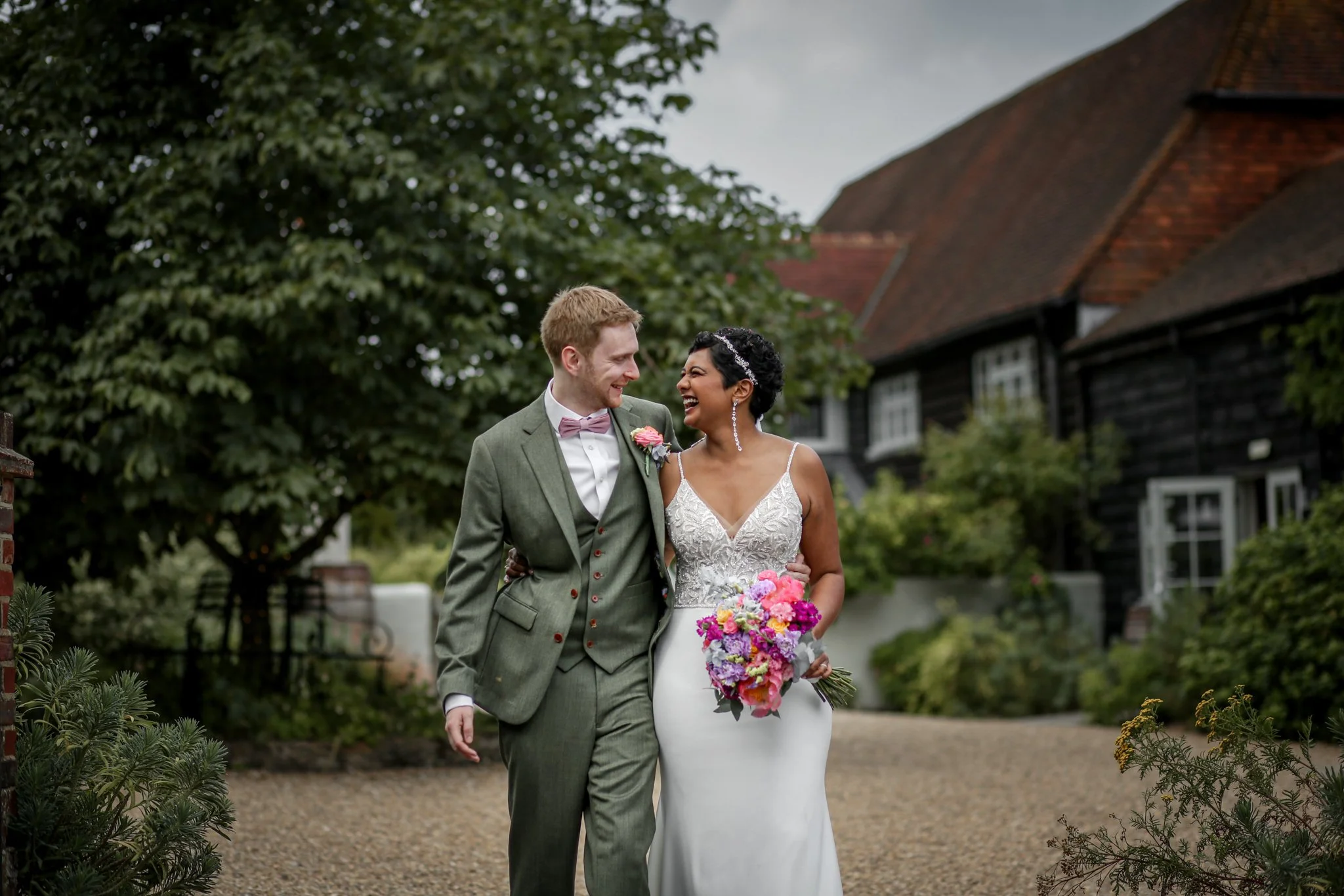 A happy couple, a man and a woman, in wedding attire walking outdoors in a garden area. The woman is holding a colorful bouquet, and both are smiling and looking at each other. The background features trees, a pathway, and a rustic house with dark wo