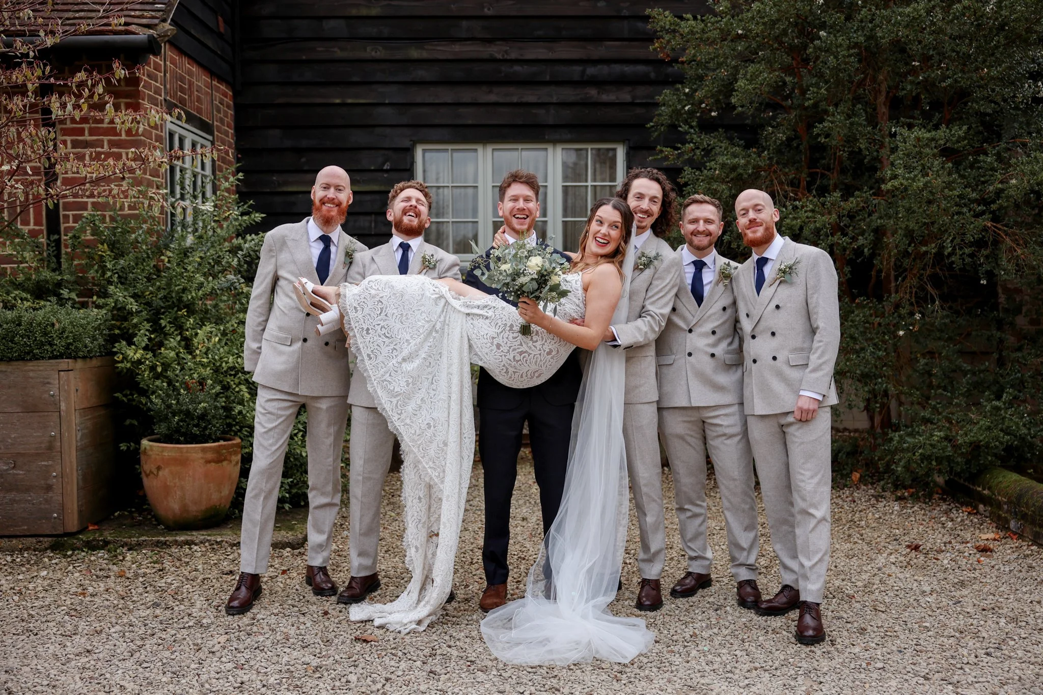 A group of seven people, six men in light gray suits with blue ties and one woman in a white wedding dress, are standing outdoors in front of a black wooden building and a brick wall. The woman is being carried by one man and holding a bouquet of flo