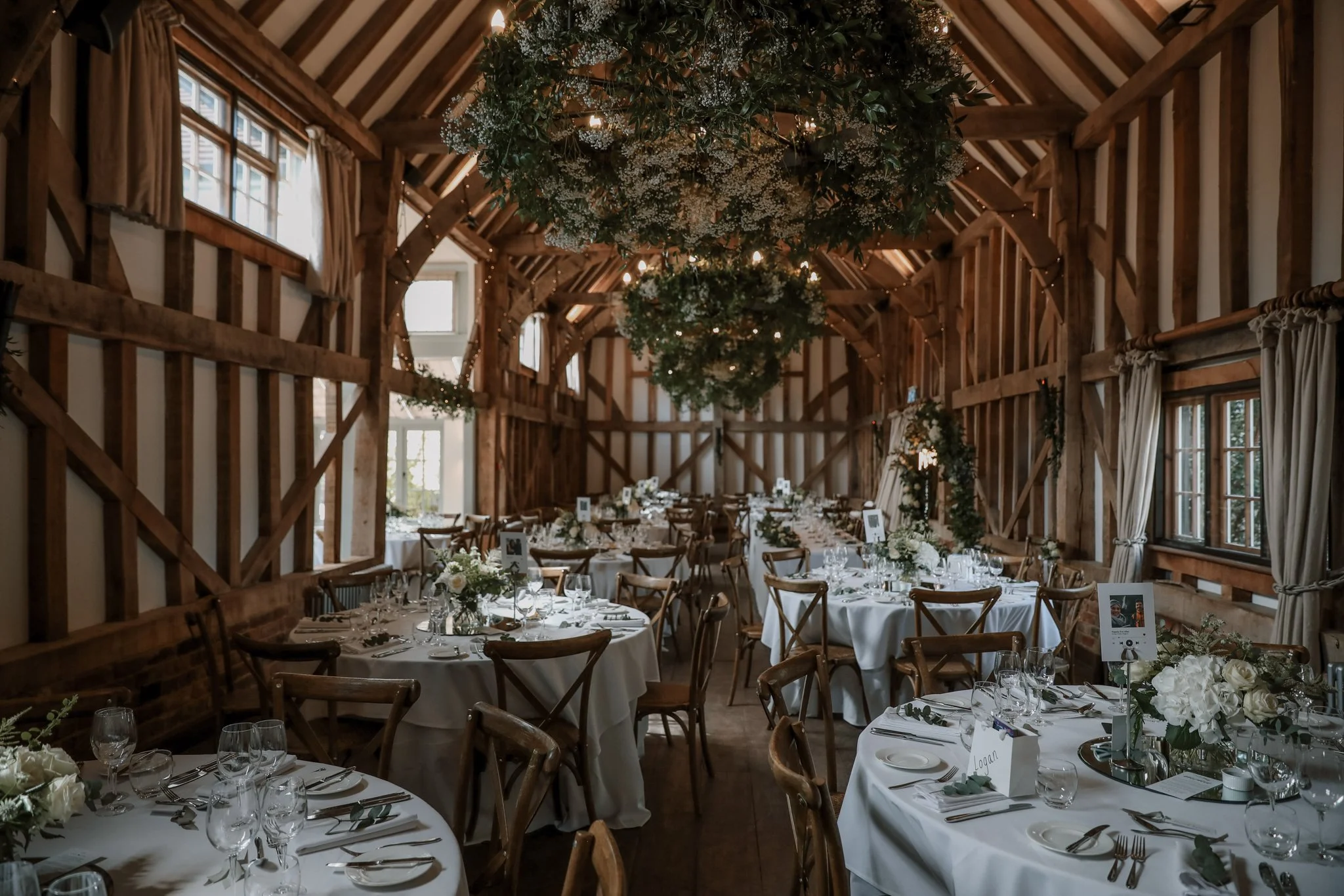 Interior of a rustic wedding reception venue with wooden beams, long tables covered with white tablecloths, floral centerpieces, and hanging greenery decorations.