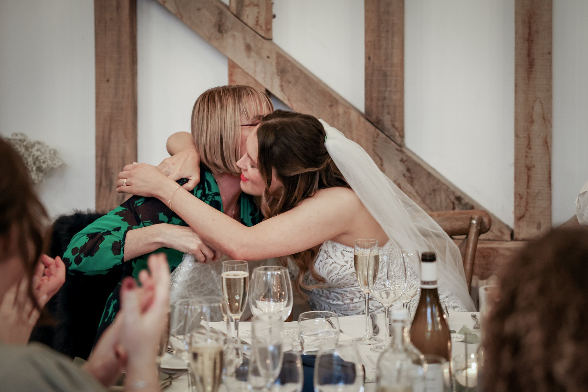 A bride and an older woman embrace and share a kiss at a wedding reception table decorated with champagne glasses and wine bottles, with wooden wall accents in the background.