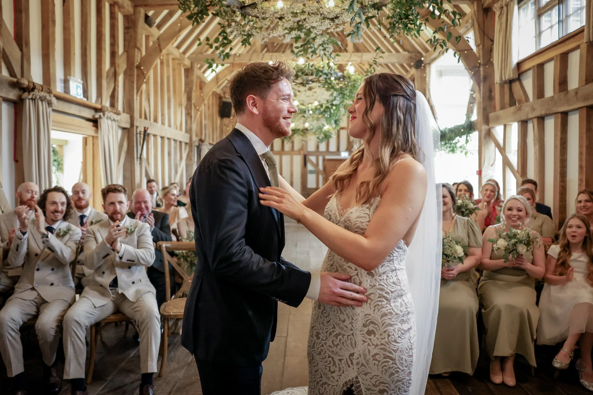 A bride and groom dancing during their wedding ceremony in a rustic barn with guests watching, smiling, and clapping.