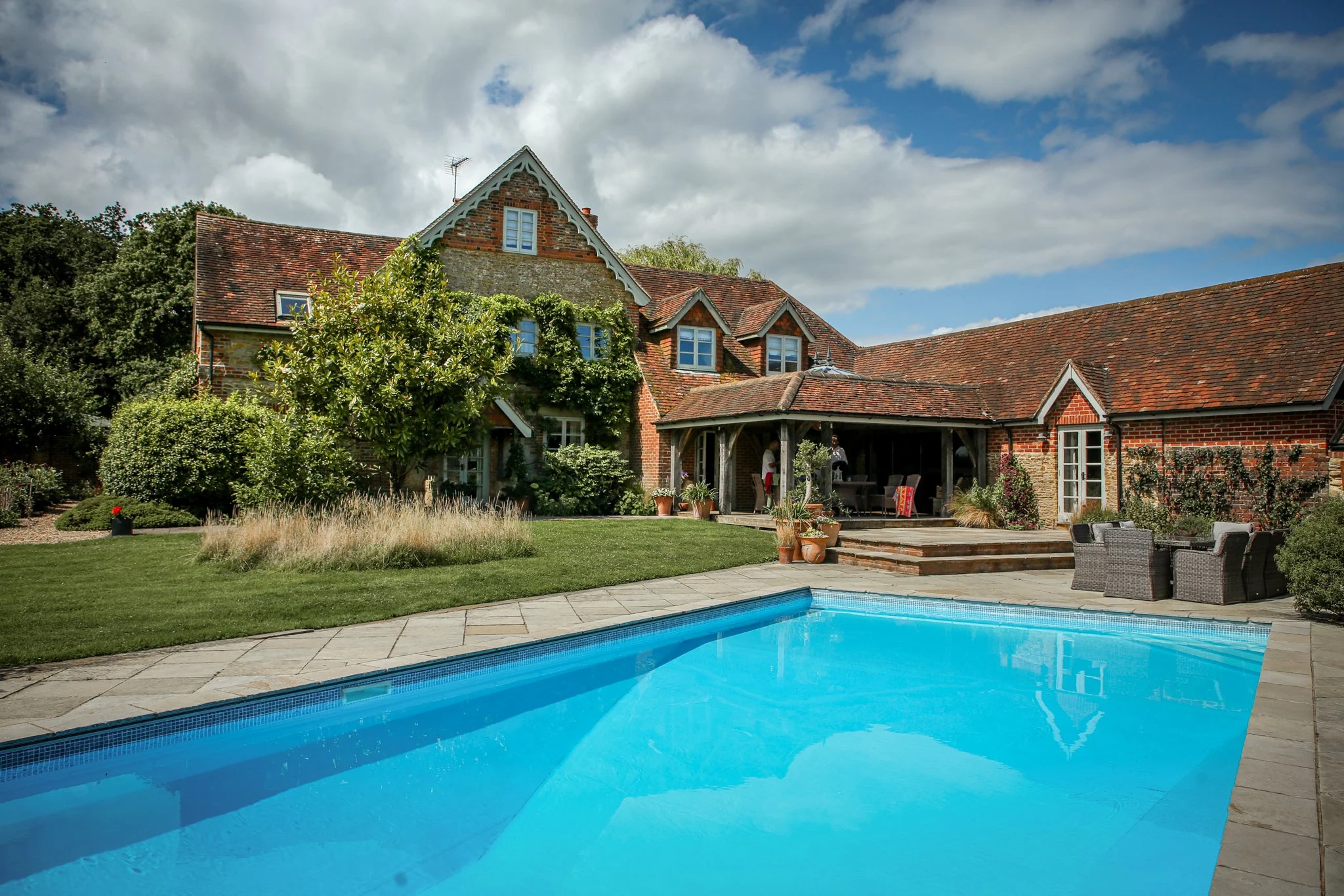 A large brick house with a steeply pitched roof and multiple windows, surrounded by trees and shrubs. In the foreground, there is a backyard with a swimming pool, a patio area with outdoor furniture, and potted plants, under a partly cloudy sky.