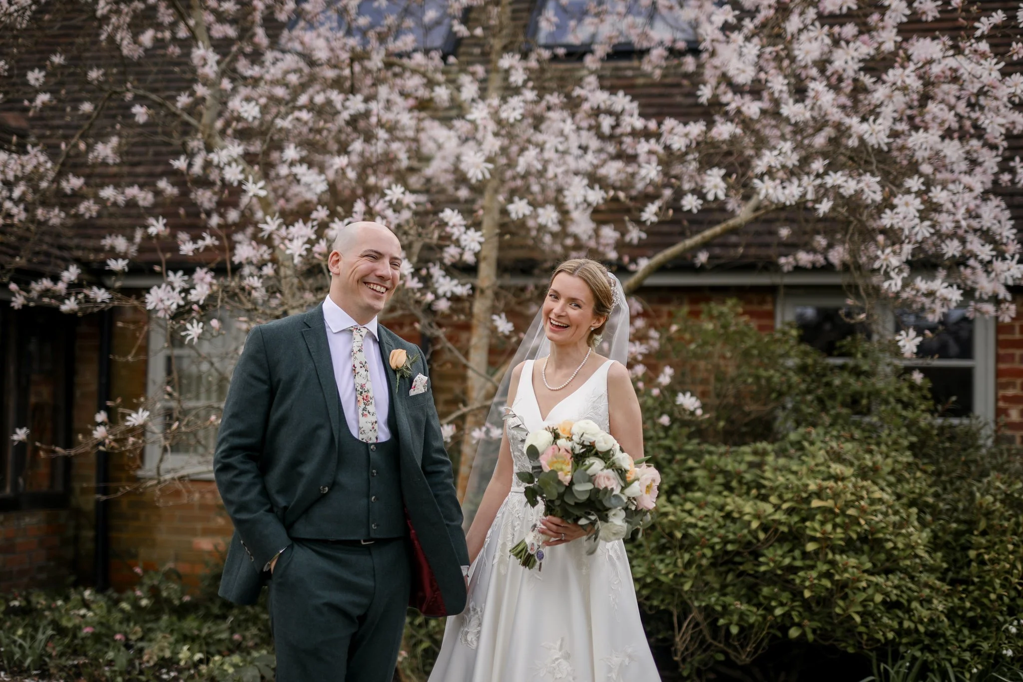 A happy bride and groom standing outdoors, smiling, with pink blossoming trees and a brick house in the background.