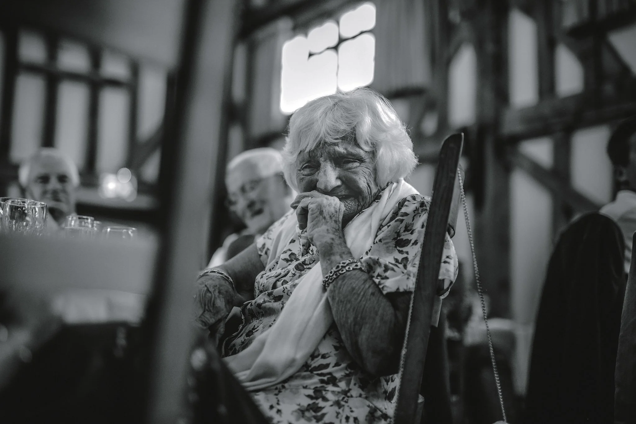 An elderly woman with white hair and a floral blouse, smiling and resting her hand on her face, seated at a table in a softly lit, rustic indoor setting with wooden beams and windows.