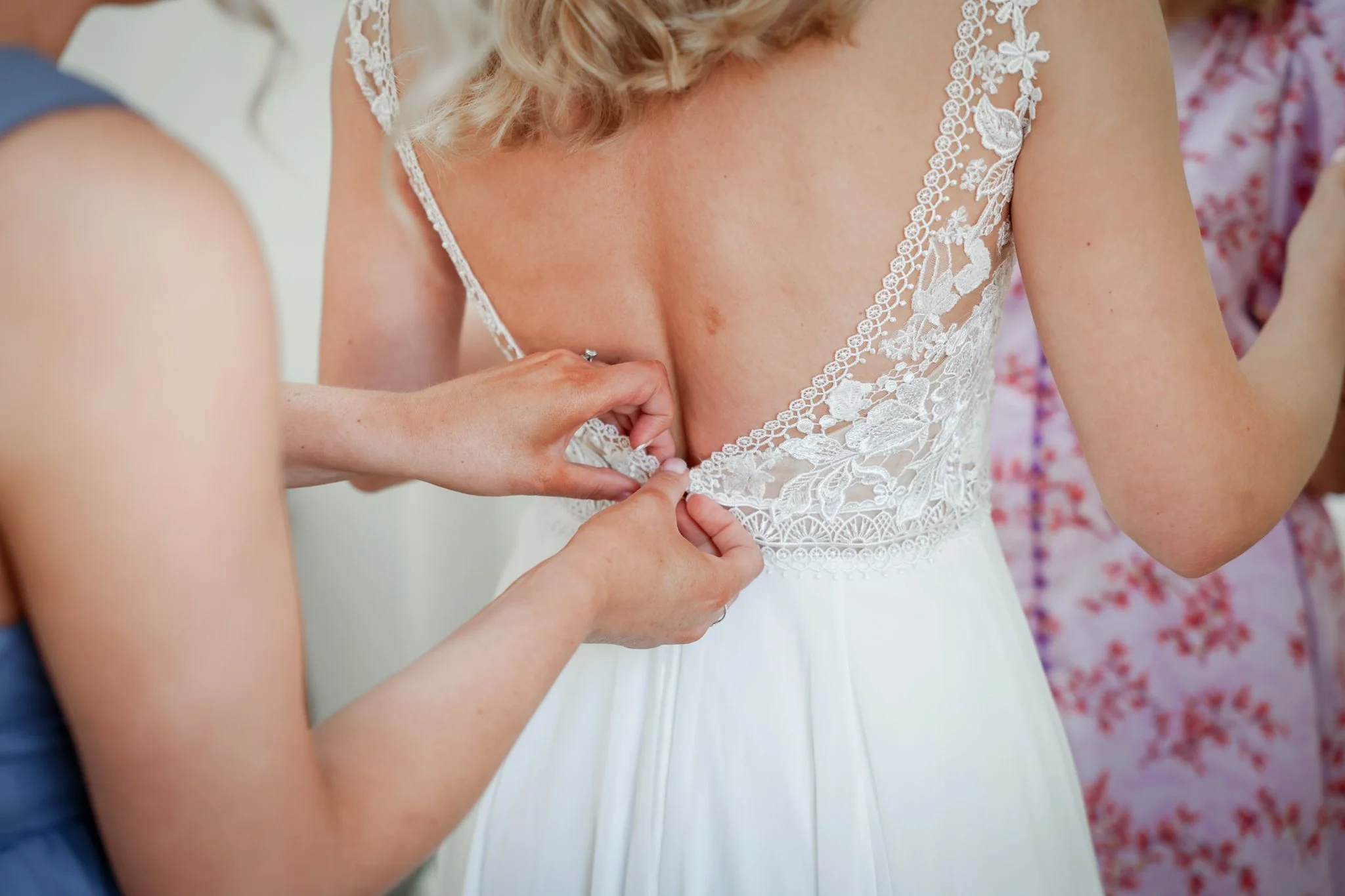 Close-up of a bride wearing a white lace wedding dress with floral embroidery, while someone adjusts or fastens the back of her dress.