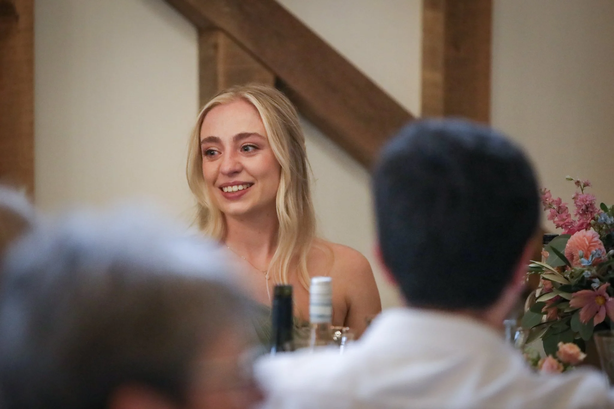 A woman with long blonde hair smiling at a gathering, with flowers on the table and glasses of drinks.