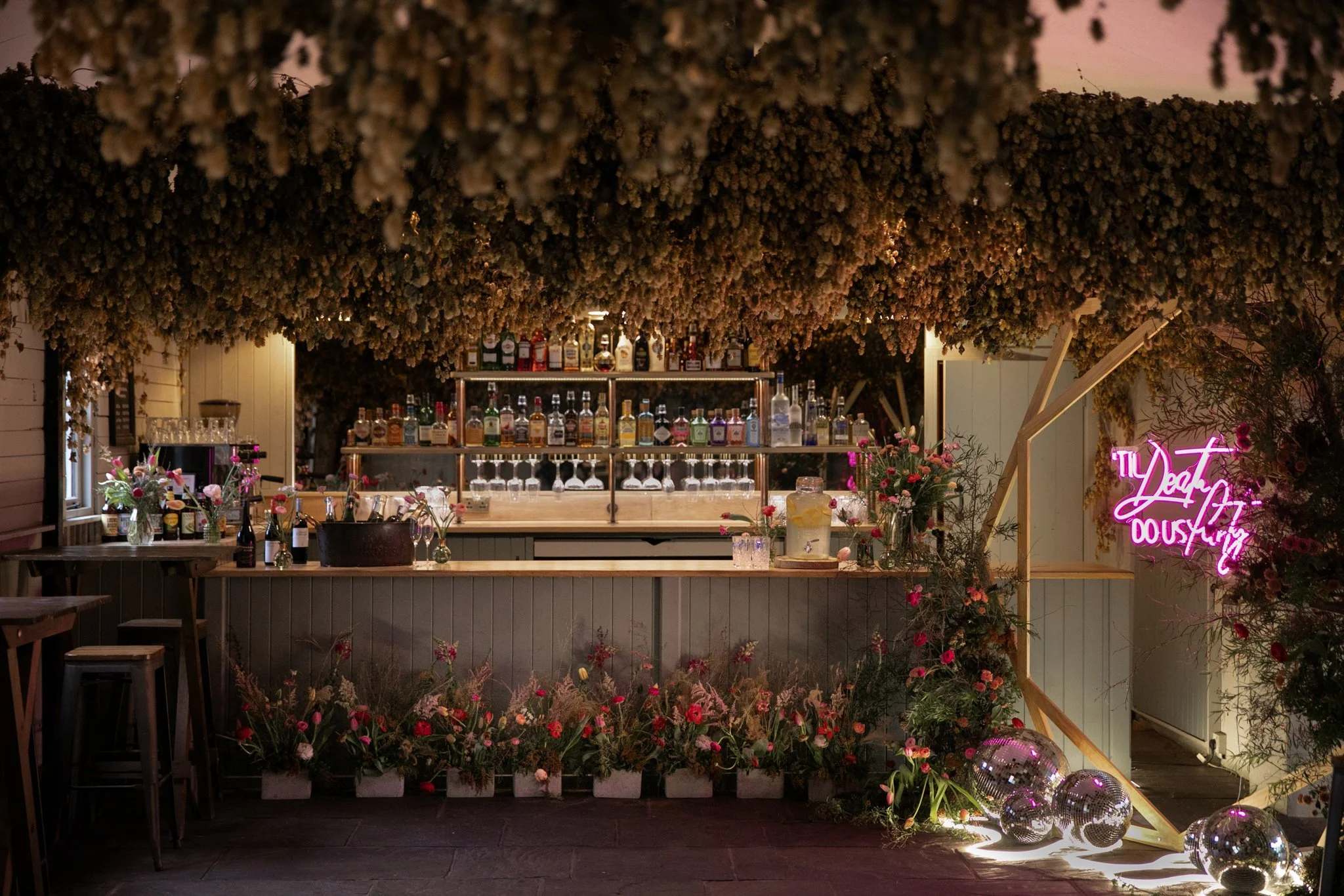A decorated bar area with a floral and lighted theme, featuring a variety of liquor bottles, wine glasses, and floral arrangements. There is a neon pink sign and reflective disco balls on the floor.