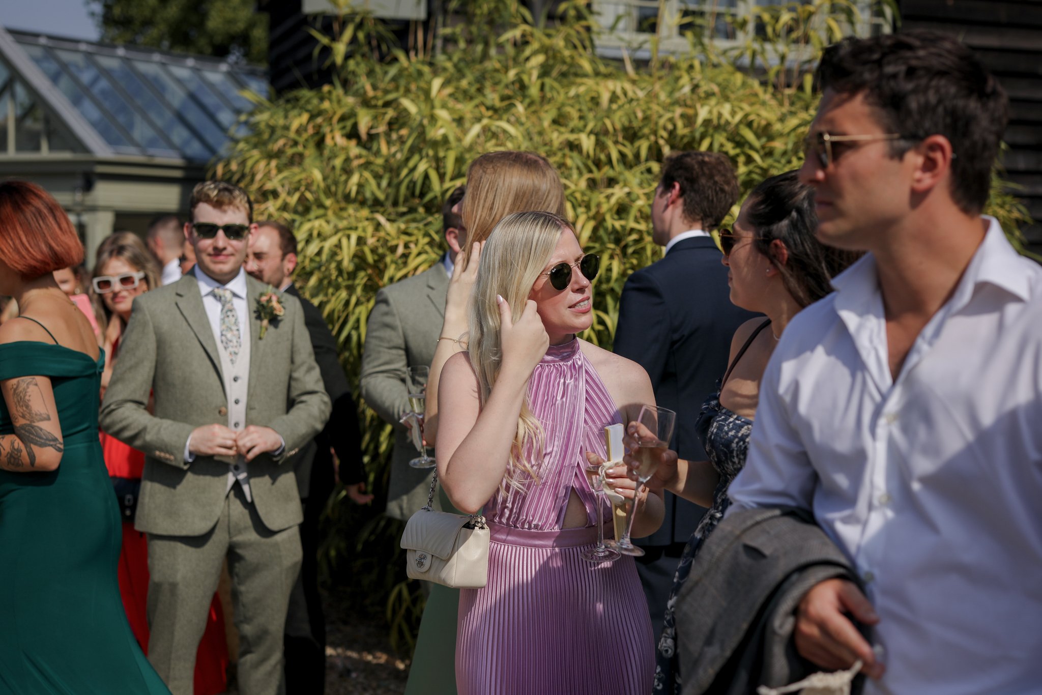 Group of people at an outdoor social event, engaged in conversation, dressed in formal and semi-formal attire, with some holding wine glasses, background features greenery and a building with a glass roof.