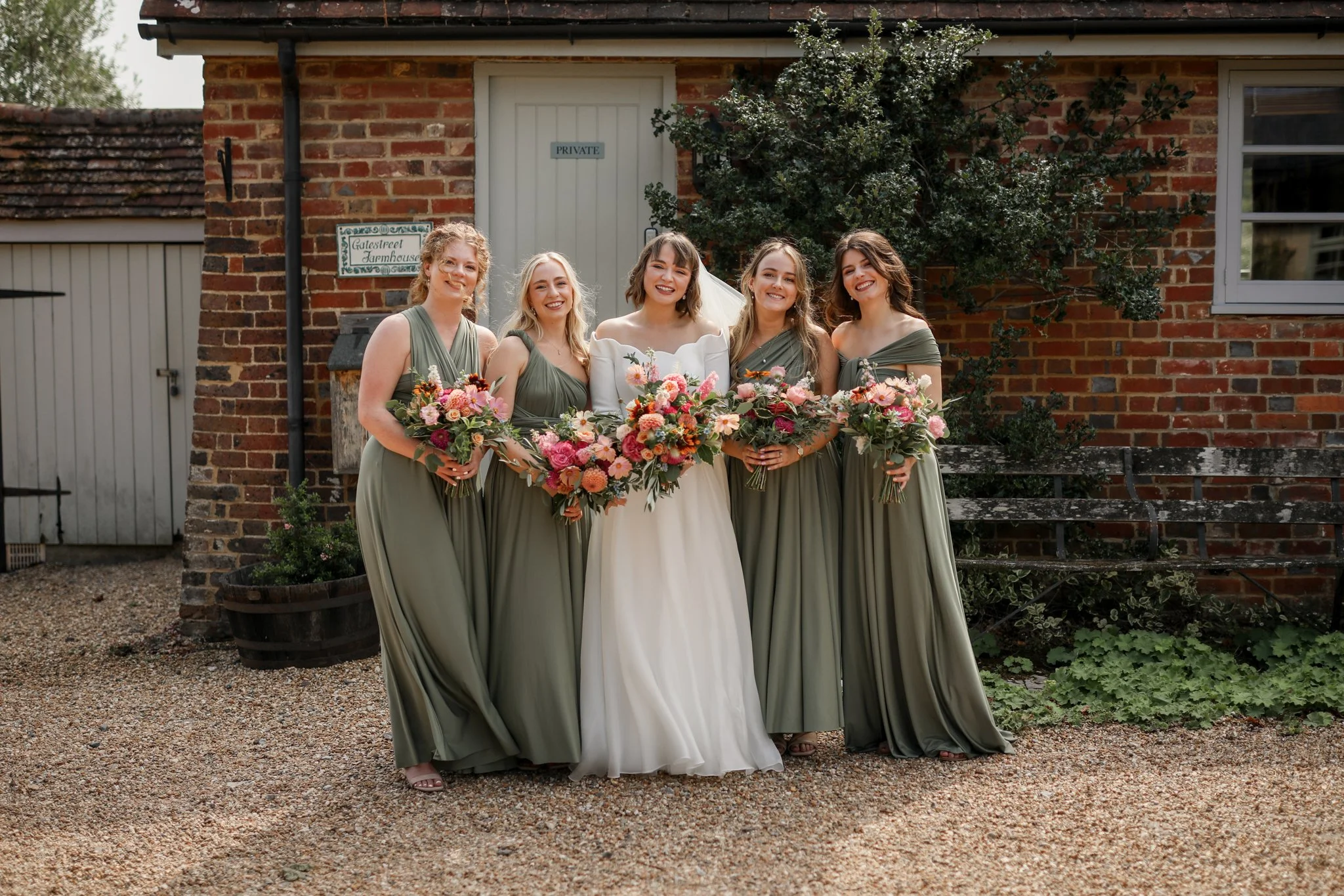 A bride in a white wedding dress and five bridesmaids in olive green dresses holding bouquets of pink and orange flowers standing outside in front of a brick building with a tree and a sign reading "Gateshead Farmhouse".