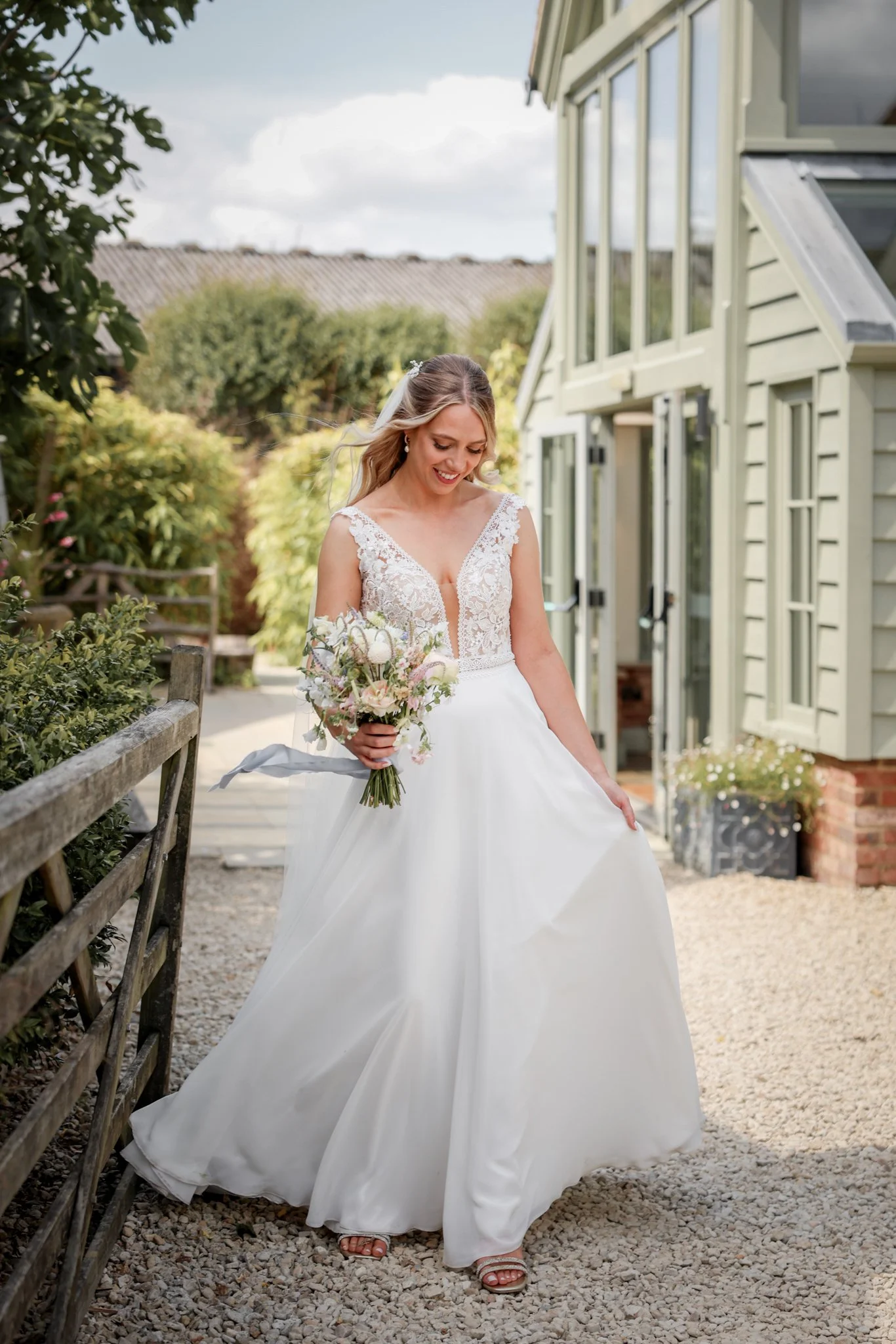 A bride in a white wedding dress holding a bouquet of flowers is walking outdoors along a gravel path. She is smiling and looking down. There are trees, a wooden fence, and a building with large windows and a sloped roof in the background.