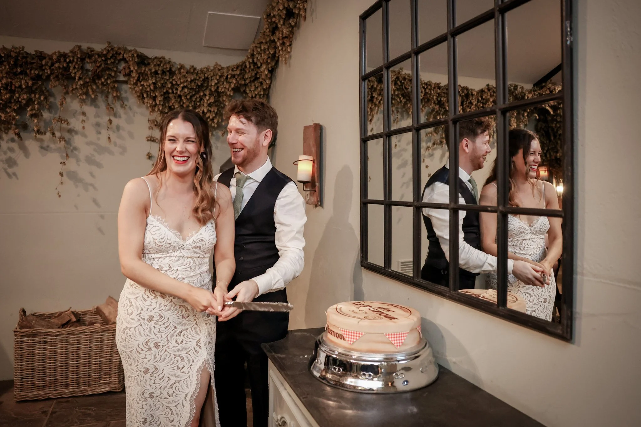 A couple, dressed in wedding attire, standing together and smiling in a warmly lit room, with a mirror reflecting their joyful expressions.