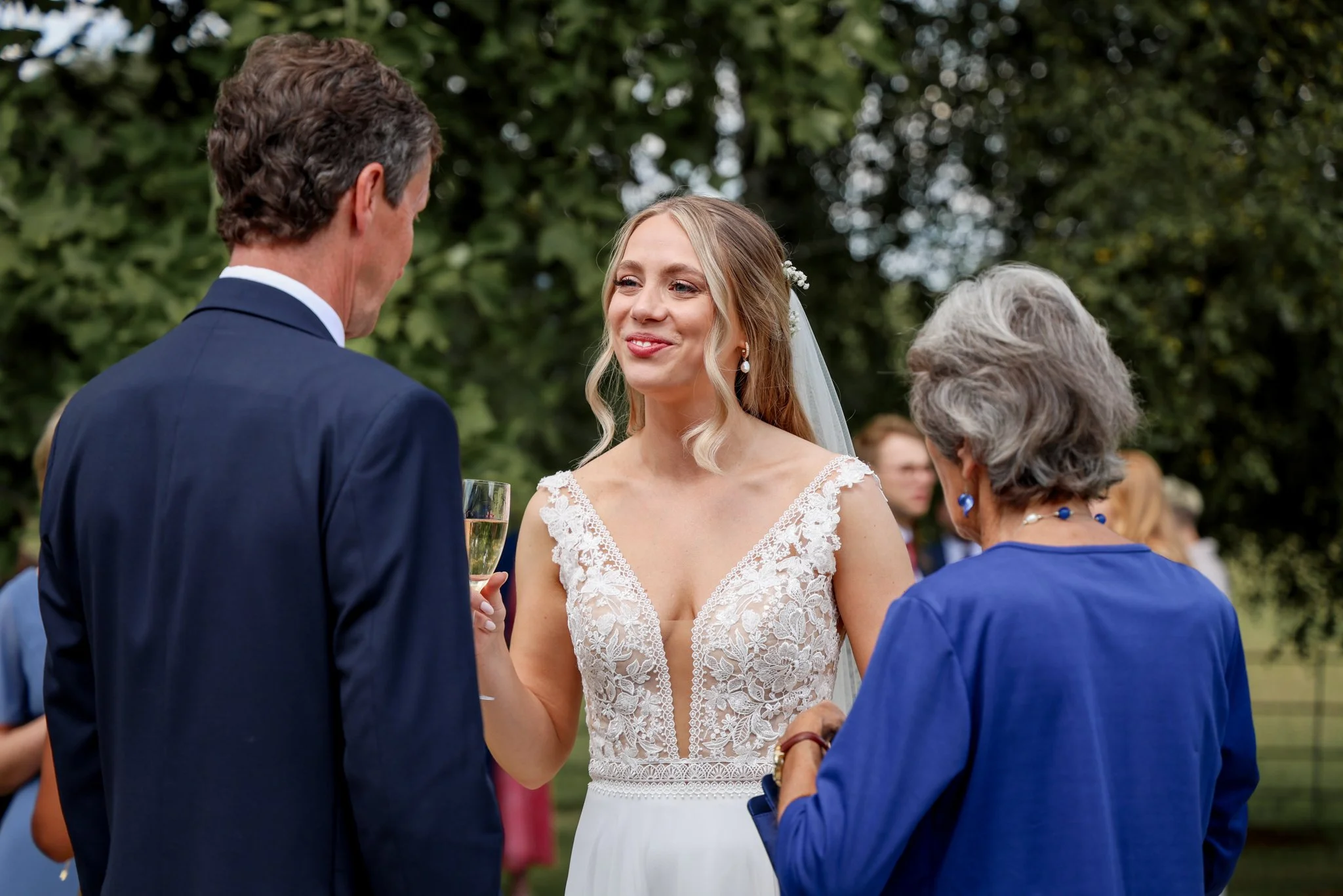 A bride in a lace wedding dress with a veil talking with a man in a dark suit during an outdoor wedding ceremony, with a woman in a blue dress nearby.