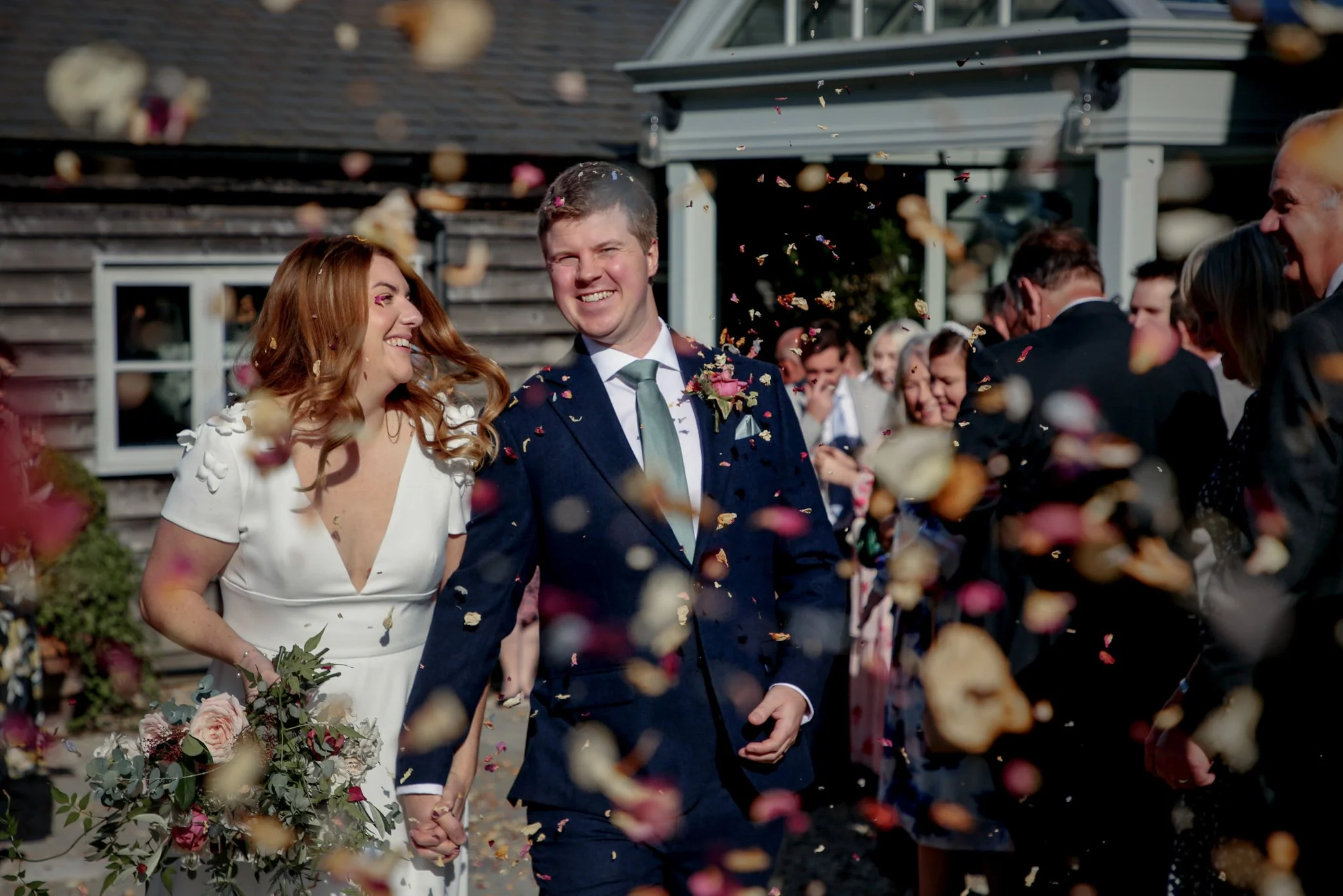A smiling bride and groom walking hand in hand at their wedding, surrounded by friends and family, with confetti falling around them outside a rustic building.