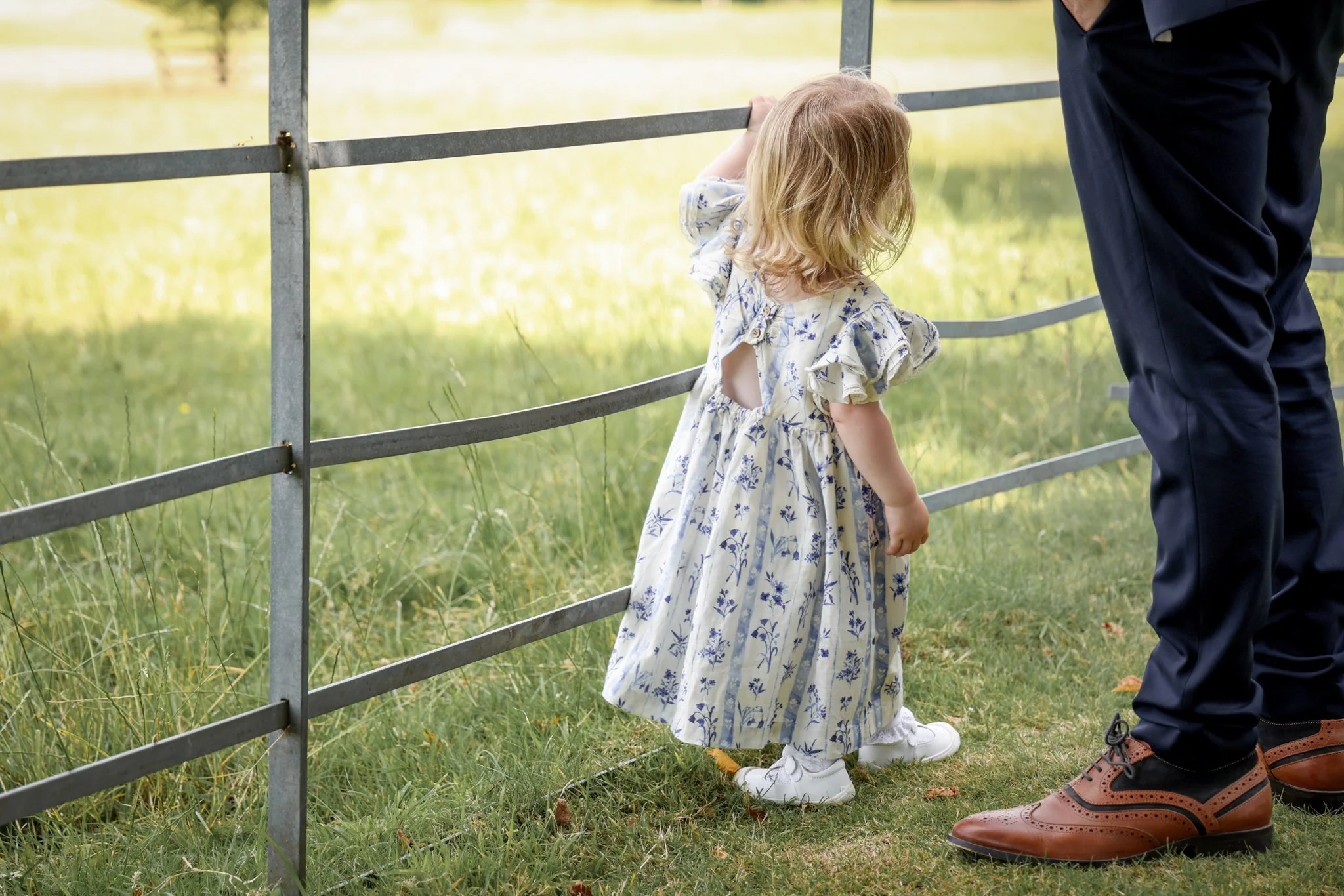 A young girl with blonde hair wearing a floral dress and white shoes stands by a metal fence, holding onto it, next to an adult man wearing dark pants and brown shoes, standing outdoors in a grassy area with trees in the background.