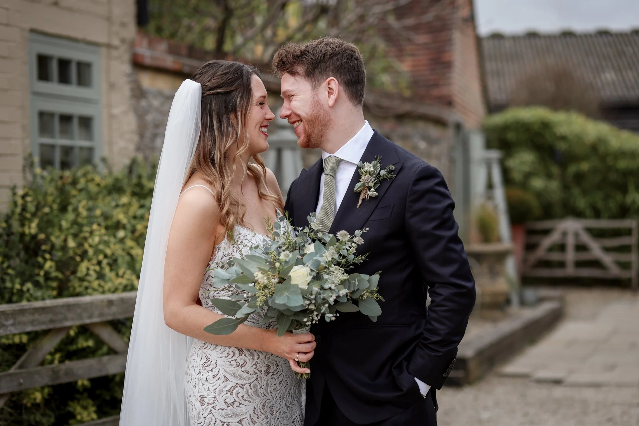 A bride and groom are standing close together outdoors, smiling and touching foreheads, with a rustic background of walls, trees, and a wooden fence. The bride is holding a bouquet of flowers and wearing a strapless lace wedding dress and a veil, whi