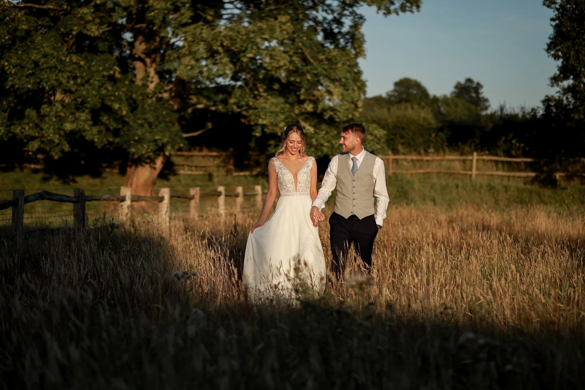 A bride and groom walking hand in hand through a field at sunset, with trees in the background.