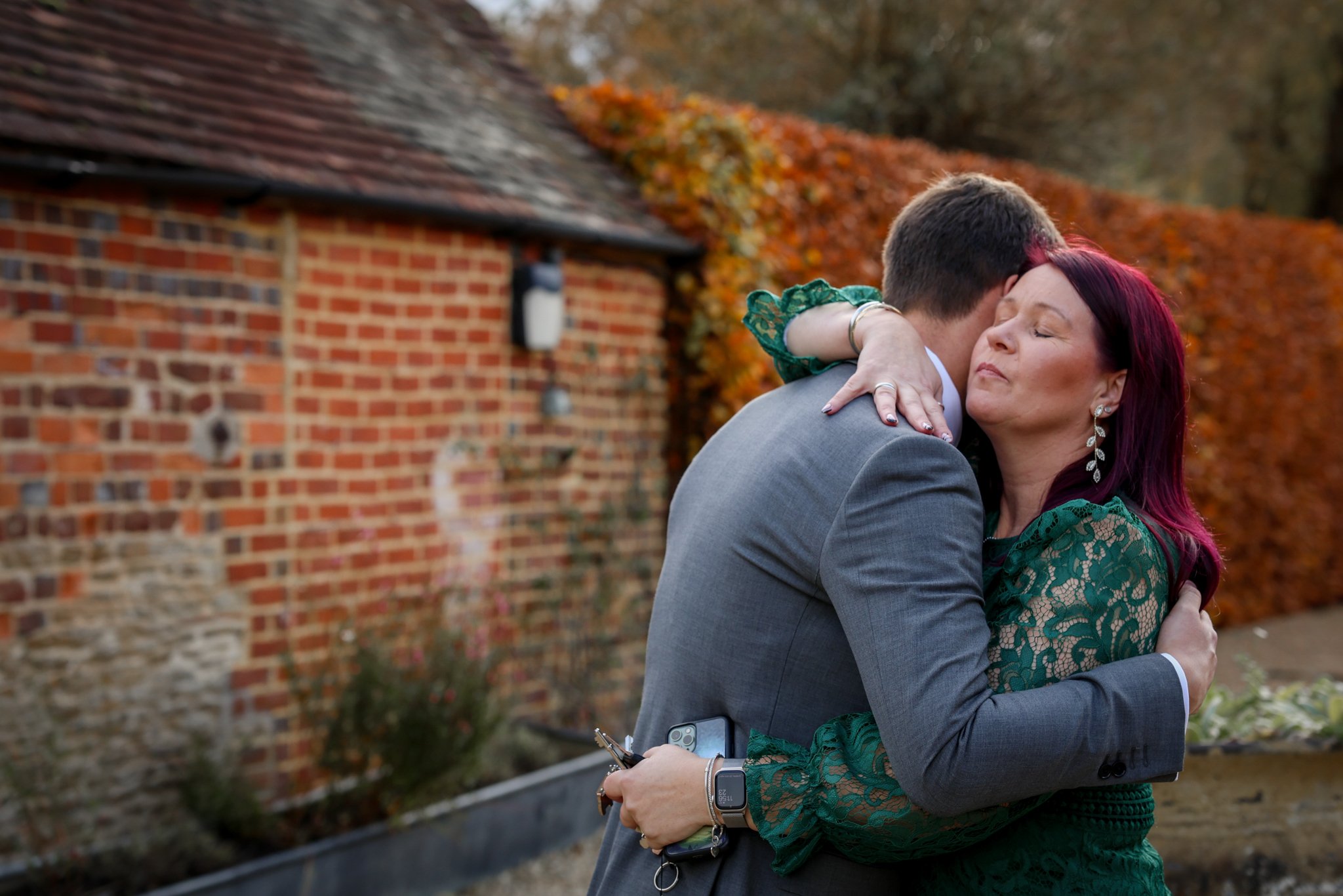 A woman with purple hair wearing earrings and a green lace dress hugging a man dressed in a gray suit holding a phone outside near a brick building with autumn foliage in the background.