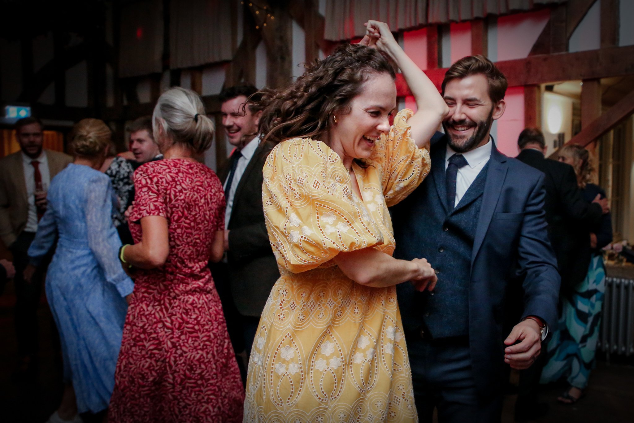 People dancing and socializing at a party, with a woman in a yellow dress dancing with a man in a blue suit, and others mingling in the background.