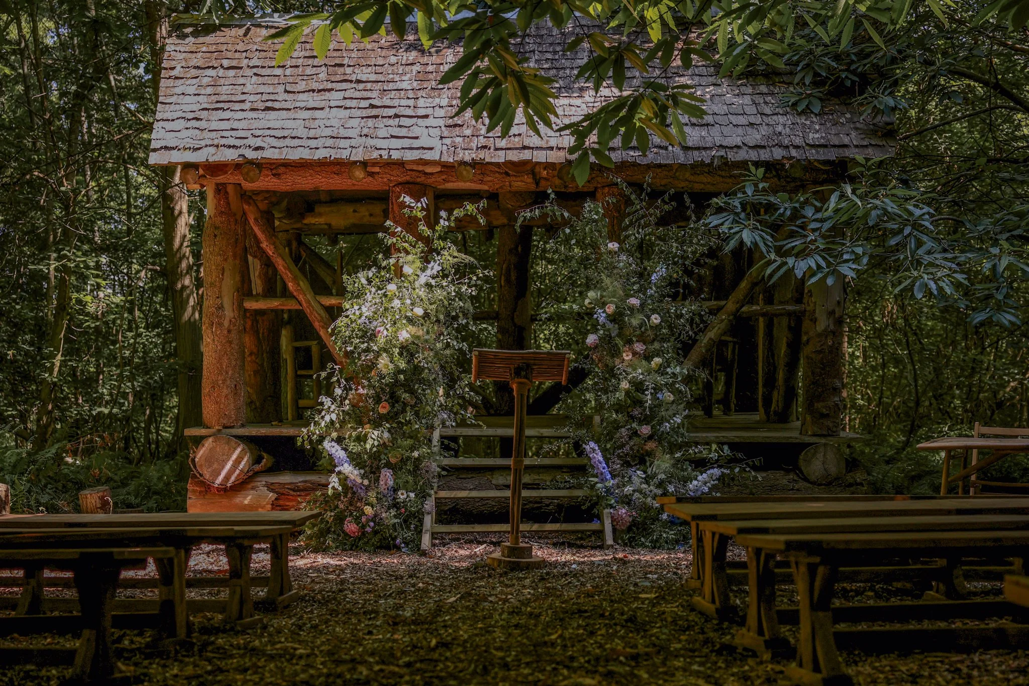 A rustic outdoor stage built into the trees in a forest, decorated with flowers, with wooden benches in front for an audience.