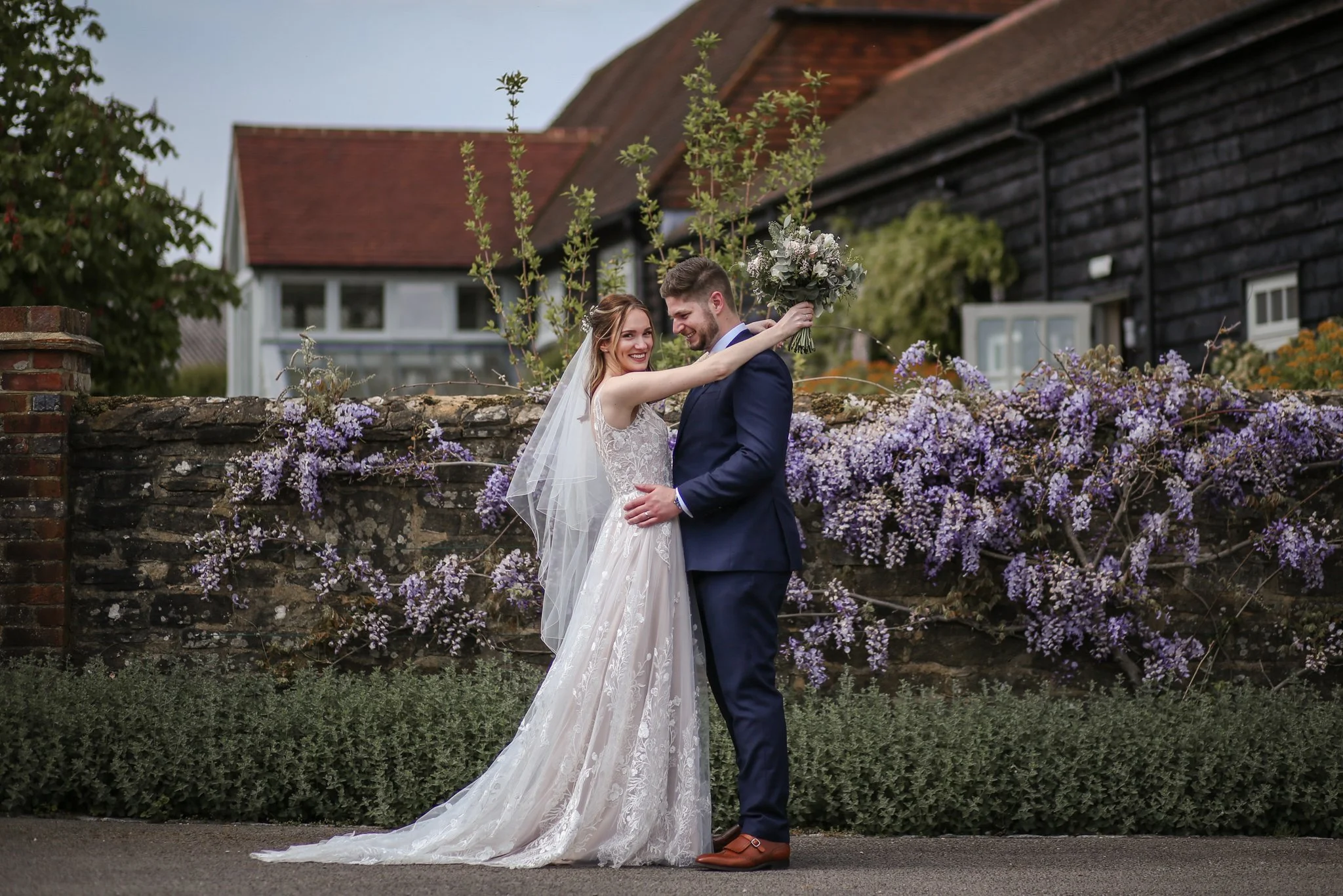A bride and groom sharing a moment outdoors in front of a stone wall decorated with purple flowers. The bride is wearing a white lace wedding gown with a veil, and the groom is dressed in a dark suit with brown shoes. The bride is holding a bouquet a